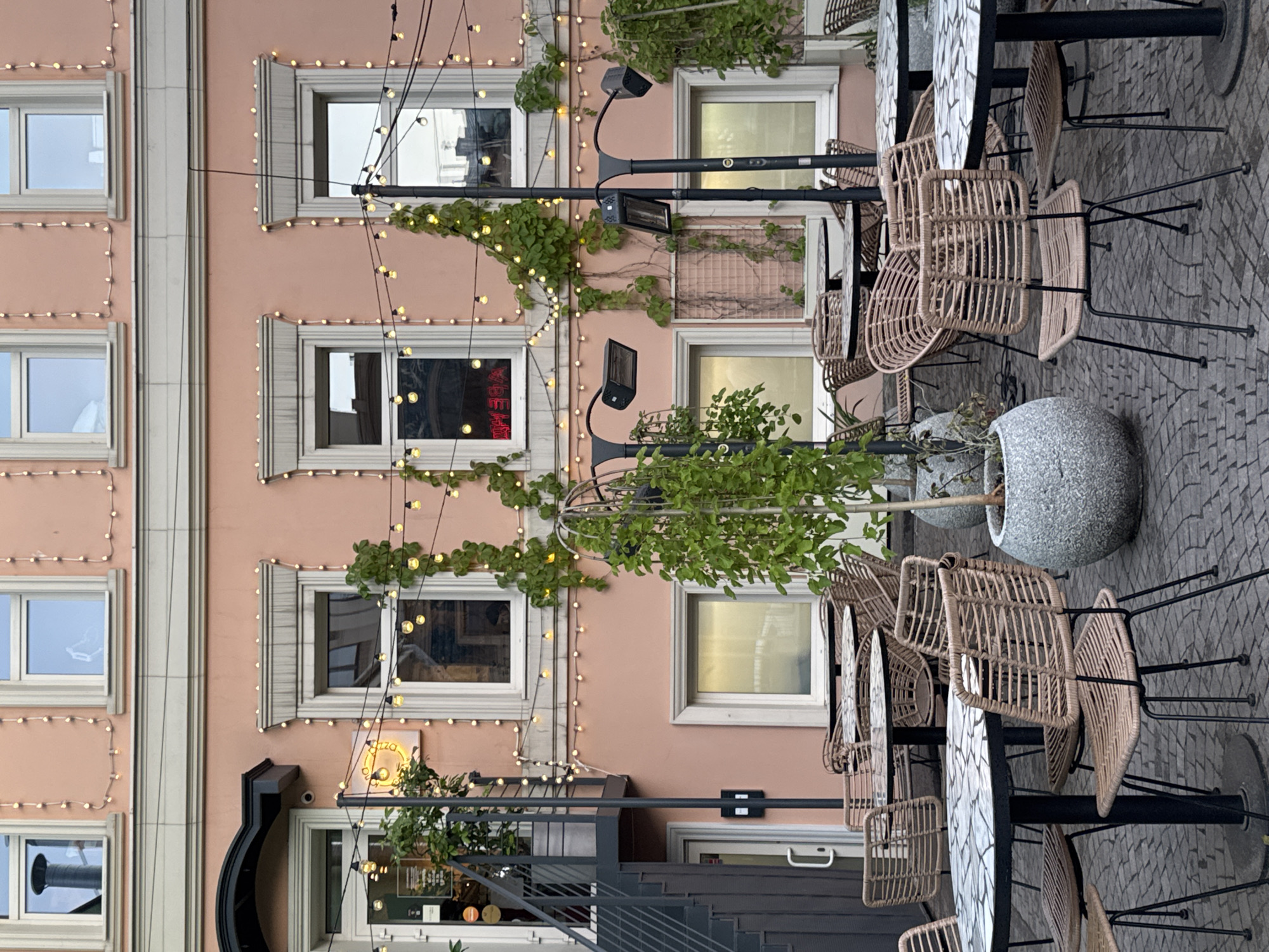 The image shows an outdoor seating area of a restaurant or cafe, likely in Nizhny Novgorod, Russia, given the prompt's context, although this cannot be definitively confirmed from visual cues alone. The scene is set against a backdrop of a pastel pink building facade. Several round tables with mosaic-patterned tops are arranged on a cobblestone patio. Wicker chairs with black metal frames surround the tables. String lights with small, warm bulbs are strung across the facade and over the seating area, creating a festive ambiance. Green vines climb the building walls, and potted plants, including a tree in a large, rough-textured stone pot, add to the greenery. Two outdoor heaters are mounted on black poles. The windows of the building are visible, some reflecting the sky and surrounding architecture. One window displays a red neon sign with reversed text, suggesting it might be a view from inside looking out, or a display for passersby. There are no people visible in the image. The overall impression is of a charming and inviting outdoor space, possibly prepared for evening service.