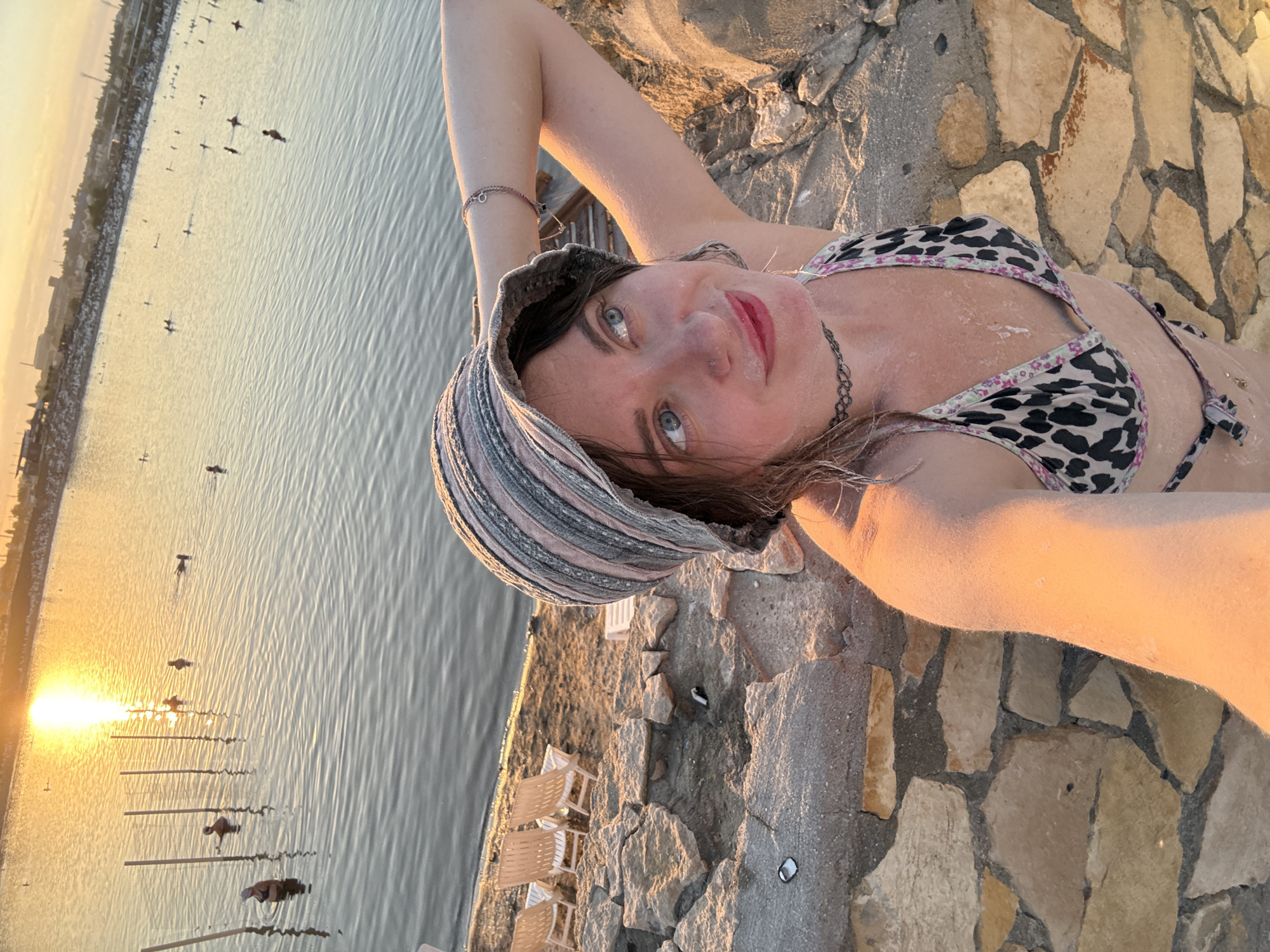 The image depicts a young woman taking a selfie in the city of Sol-Iletsk, Russia, near a body of water. The woman is wearing a bucket hat and a leopard-print bikini. She has light skin and blue eyes and is looking to her right with a slight smile. Her left arm is raised behind her head. In the background, the sun is setting over the water, casting a warm glow. Several people are visible in the water, some swimming and others standing. There are also several beach chairs on the shore to the left of the frame. The foreground shows a stone wall. The overall scene suggests a leisurely day at a resort or a natural body of water.