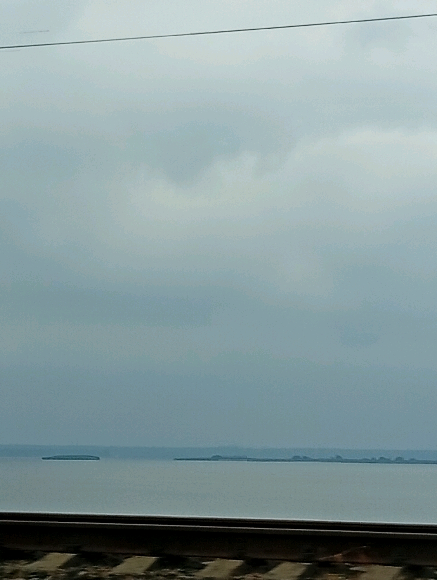 The image captures a landscape scene from the perspective of a moving vehicle, likely a train, as indicated by the presence of train tracks in the foreground. The view is dominated by a vast, calm body of water under an overcast sky. Several small, elongated islands or land masses are visible in the distance, scattered across the water. The overall atmosphere is muted and somewhat somber due to the cloudy weather. The image is a still from an unknown location in an unknown city.