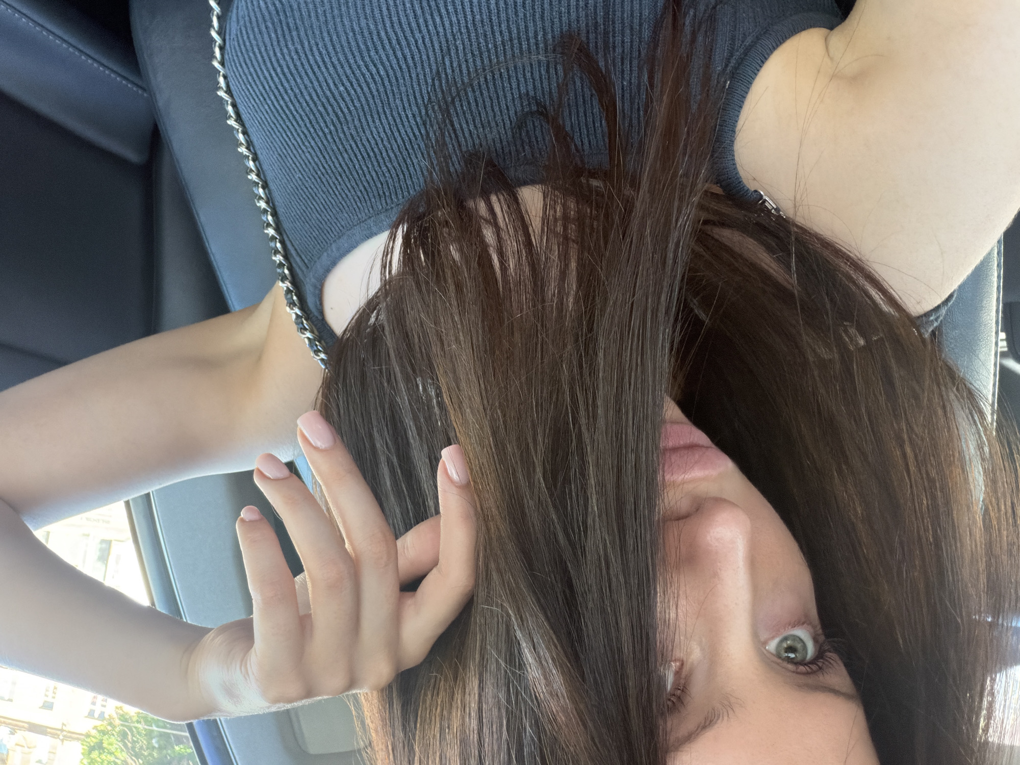 This media file is a close-up, vertically oriented photograph of a woman in what appears to be the interior of a car. The woman has long, dark brown hair that partially obscures her face. Her right eye is visible and appears to be green or hazel, with a wide-open gaze. Her lips are slightly parted. Her left hand, with neatly manicured pale pink nails, is raised and touching her hair, with her fingers delicately posed. She is wearing a dark grey ribbed tank top with a silver chain strap visible over her shoulder. The background shows the dark leather interior of a car, likely a seat. Sunlight is streaming in, suggesting it is daytime. The presence of buildings and trees visible through a car window in the background indicates an urban or suburban outdoor setting. The overall mood conveyed is casual and personal, possibly a selfie taken during a car ride. No visible text is present in the image. The location context is Zaporizhzhia, Ukraine.