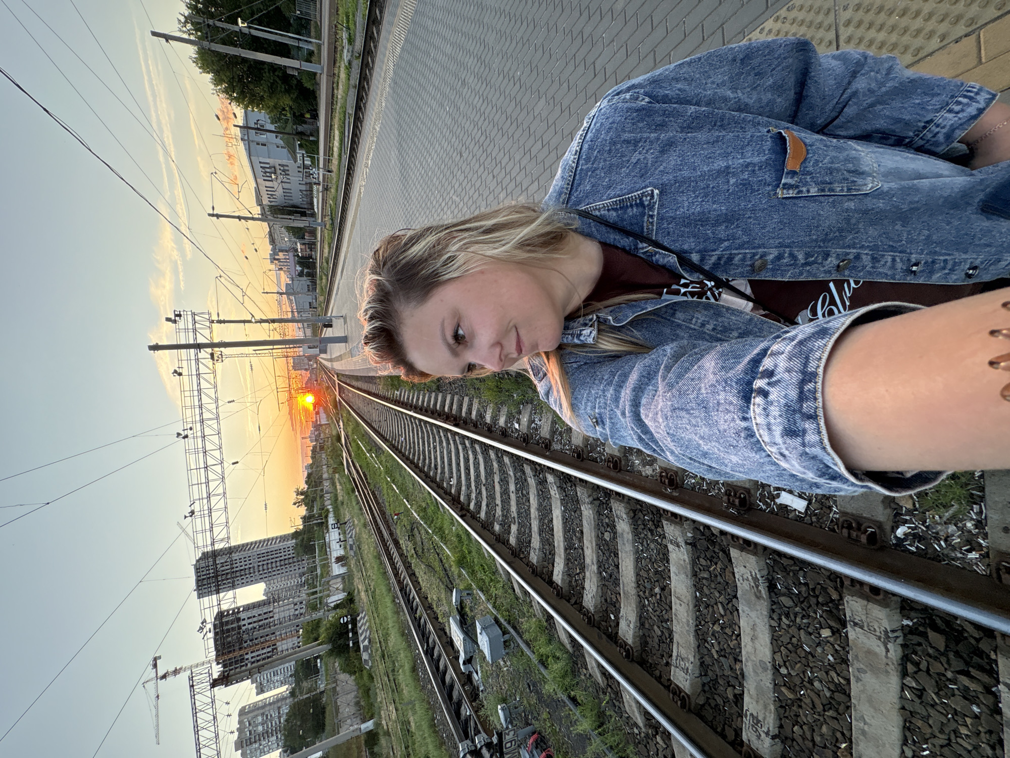 An outdoor, eye-level, medium shot captures a young woman from the chest up, posing on a train platform during a sunset in Volgograd, Russia. She is looking to her right, with her blonde hair slightly windswept and her face turned away from the camera. The woman is wearing a denim jacket over a maroon t-shirt. In the background, train tracks stretch into the distance, with overhead power lines crisscrossing the sky. The sun is setting, casting a warm glow and creating an orange hue in the sky. The platform is paved with interlocking bricks, and there are buildings and construction cranes visible on the horizon. The scene conveys a sense of urban transit against a natural, fleeting moment.
