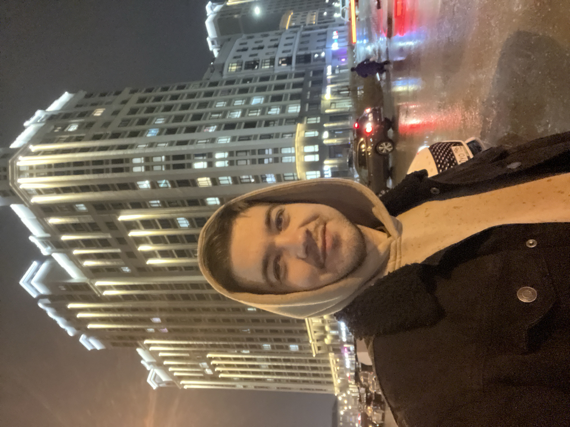 A nighttime selfie of a man wearing a hooded sweatshirt and a dark coat with visible buttons. He's positioned in front of a large, brightly lit building with many windows, likely an apartment complex or office building. Streetlights and reflections on wet pavement suggest it has recently rained or is currently raining. In the background, there are cars and other pedestrians. The location appears to be in a city. Based on the architecture and overall appearance of the image, the location may be in Yekaterinburg, Russia. The building is identifiable as the "Iset Tower", a notable skyscraper in the city.
