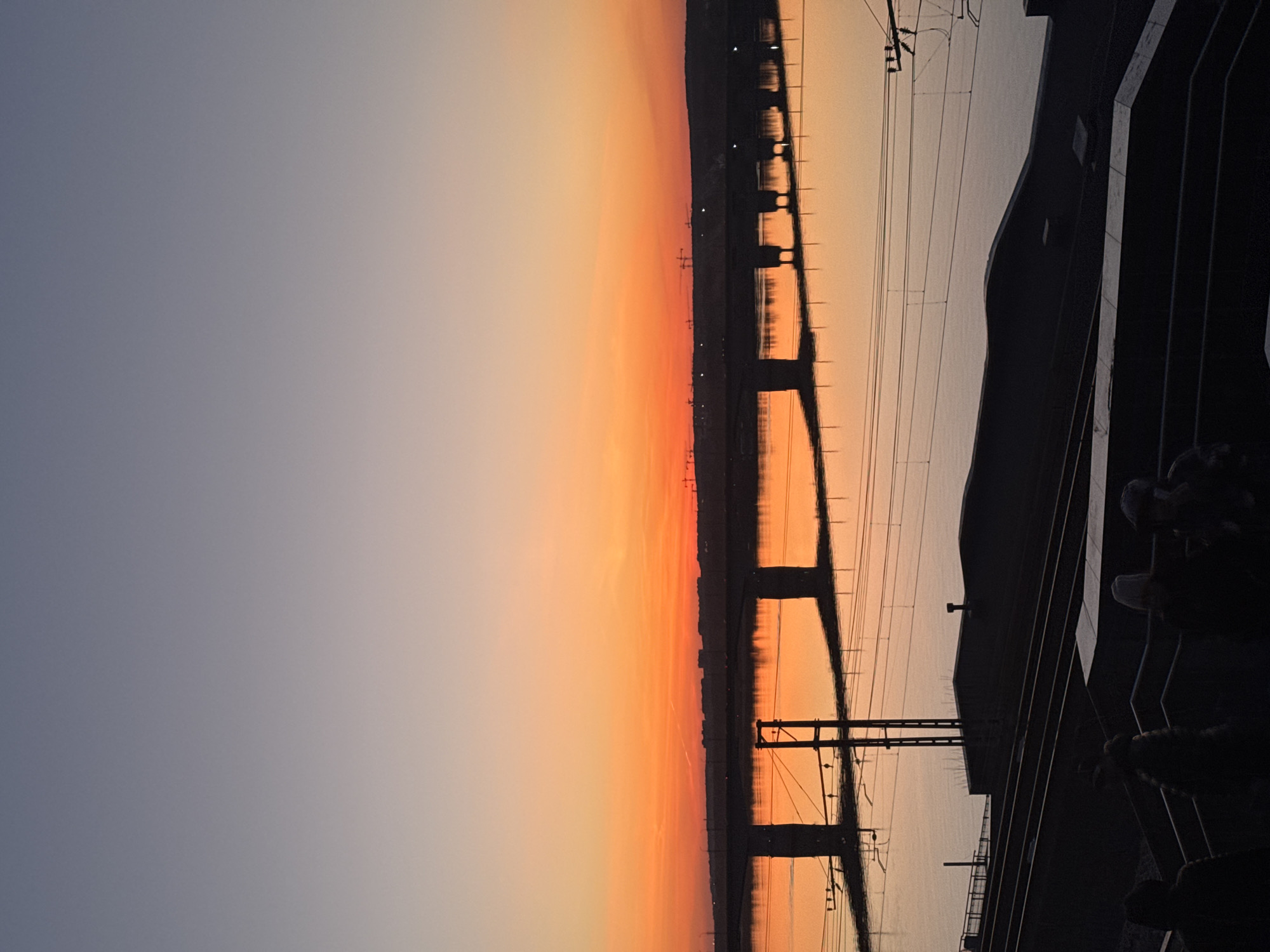 The image depicts a sunset view in Perm, Russia. The sky transitions from a pale blue-gray to a vibrant orange near the horizon. A bridge-like structure spans the scene, its support pillars reflected in the water below. Power lines and the silhouette of a building with a railing are visible on the right side of the frame, possibly a train or a river embankment. Details about people are not visible.