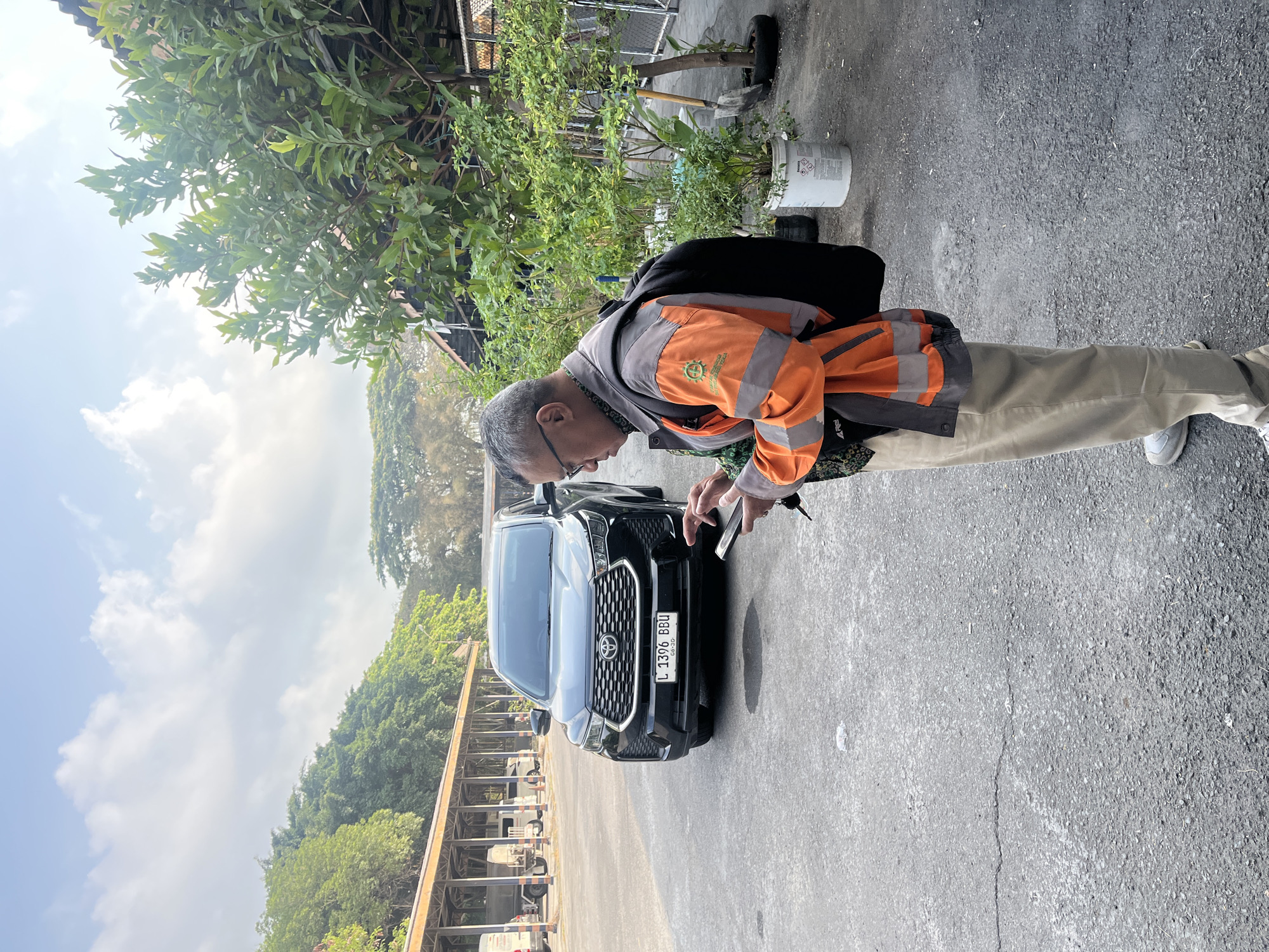 A man wearing an orange and grey high-visibility jacket over a patterned shirt and beige trousers is standing in an outdoor parking lot and looking at his phone. He has a black backpack on. To his left, a black Toyota Innova is parked, with its license plate reading "L 1396 BBU". The background shows a covered parking area with other vehicles, trees, and a cloudy sky.