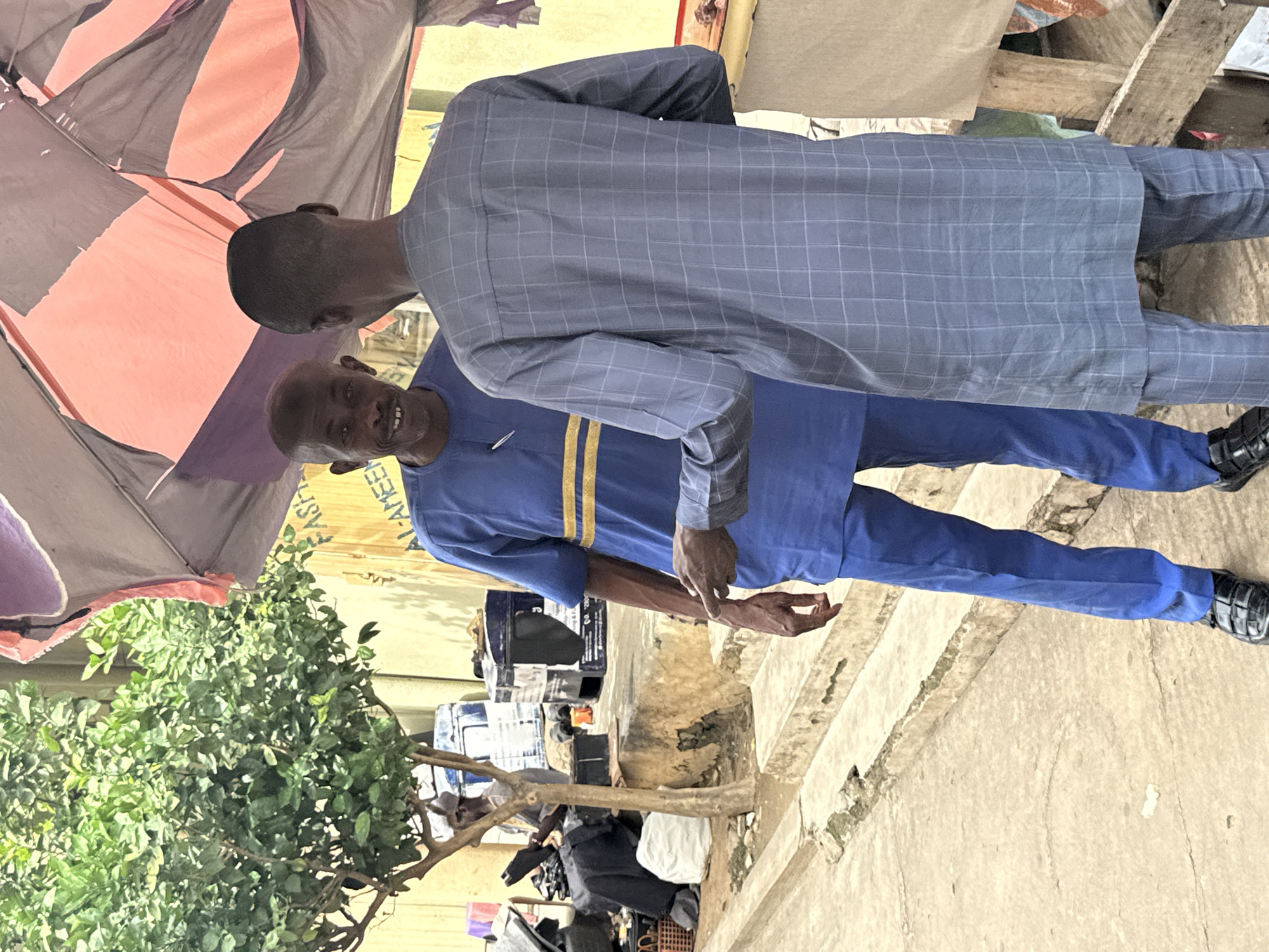 Two men are standing under a torn, colorful canopy in Abuja, Nigeria. The man on the left is wearing a blue tunic with yellow stripes and black sandals. He is smiling and pointing his finger towards the man on the right. The man on the right is wearing a blue checkered tunic and matching pants. He is facing away from the camera, looking down. In the background, there is a small tree, some stairs, and various objects including boxes and baskets.