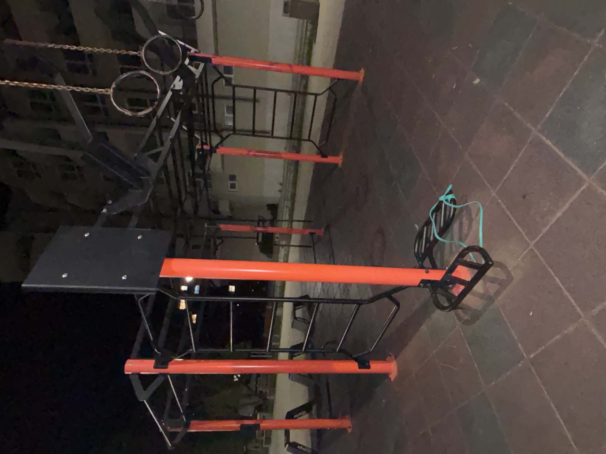 The image shows an outdoor fitness structure with metal bars and platforms in Burgas, Bulgaria. The structure is primarily composed of black metal framing with orange horizontal bars. It includes features like monkey bars, rings hanging from chains, and a raised platform. The structure sits on a paved surface with a tile pattern. The background shows a building with windows. The lighting suggests it is dusk or night. There are no people visible in the frame.