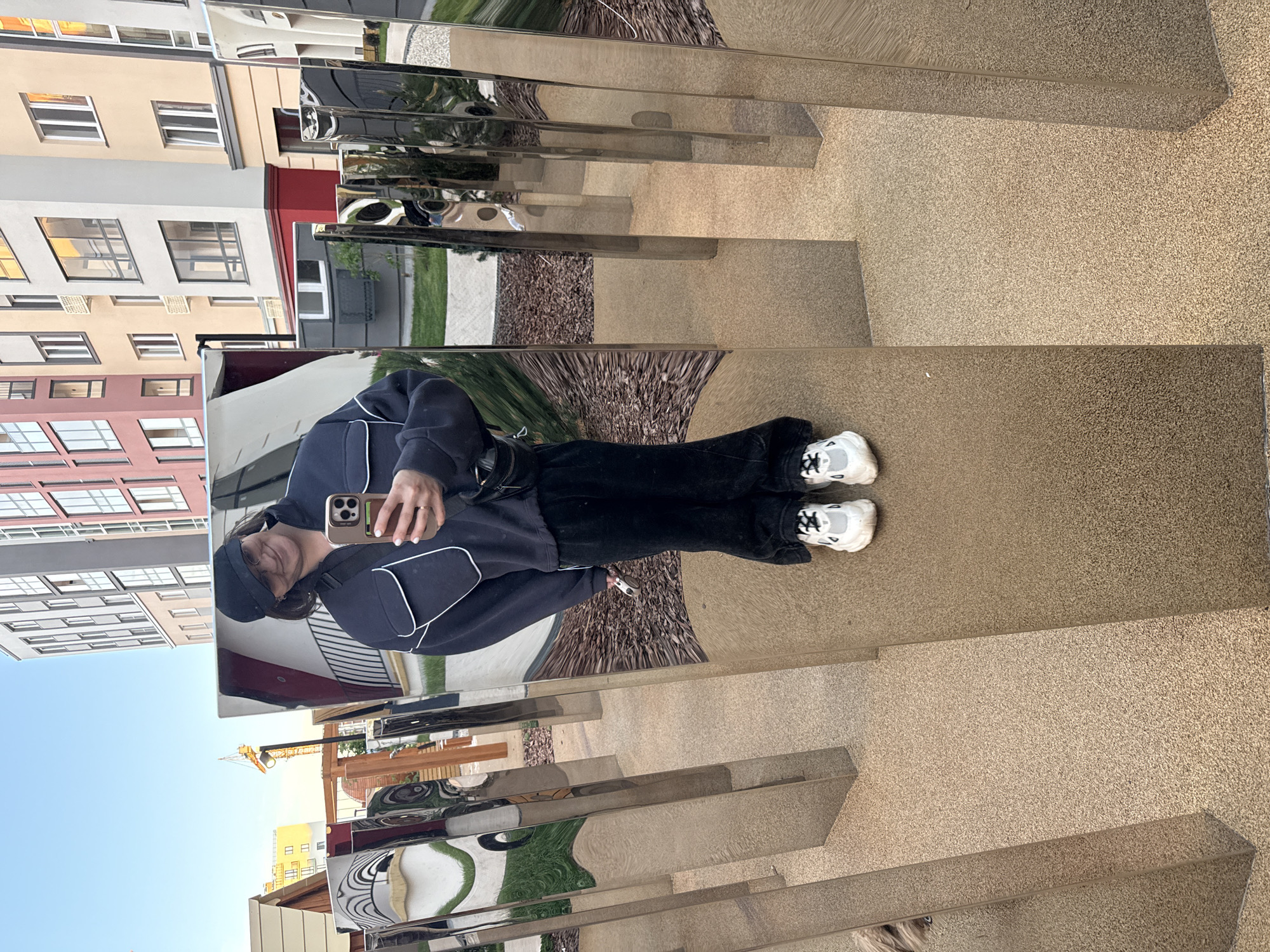 A person is captured in a selfie reflected in a series of tall, rectangular mirrored panels. The image appears to have been taken in a public outdoor space, likely an urban or residential area in Pervomaysky, Russia.
The individual, dressed in a dark blue or black top with white piping, black pants, and white athletic shoes, is holding a light-colored smartphone to take the photo. A black cap is worn, and a ring is visible on the person's left ring finger. The mirrored panels create distorted reflections of the surroundings, including portions of modern residential buildings with facades in shades of beige, white, red, and pinkish-brown. These buildings feature numerous windows.
The ground surface is a light brown, speckled material, possibly asphalt or a rubberized playground surface. Reflections also show patches of green grass and areas covered with dark wood chips. Above the buildings, a clear blue sky is visible, suggesting good weather during daytime, possibly late afternoon or early evening given the soft light. A construction crane is faintly reflected in the distance. No legible text is visible in the image.