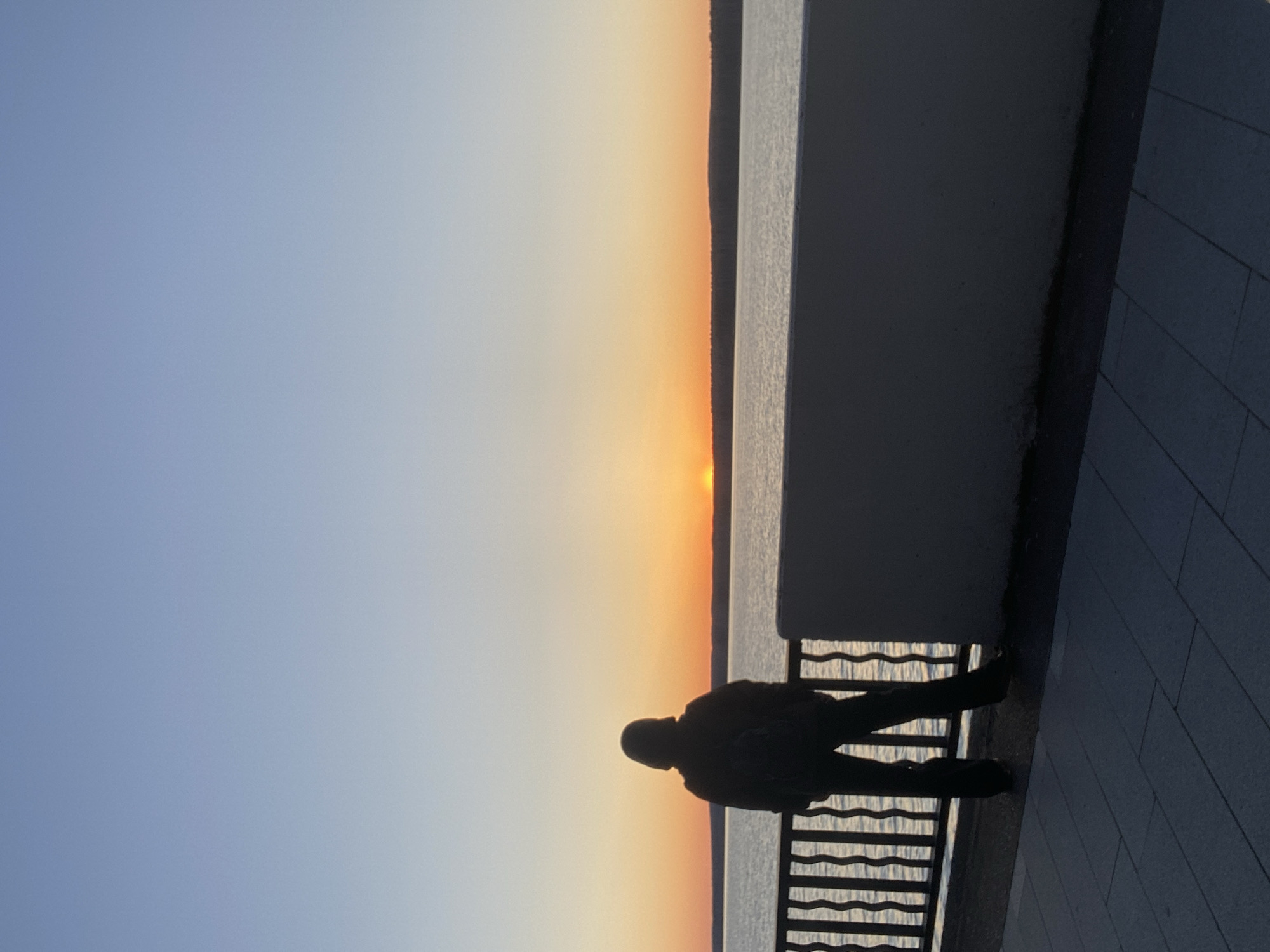A person stands silhouetted against a sunset over a body of water. They are leaning against a railing. A gray wall is on the right side of the image, and the ground is paved with gray tiles. The location is Chaikovsky, Russia.