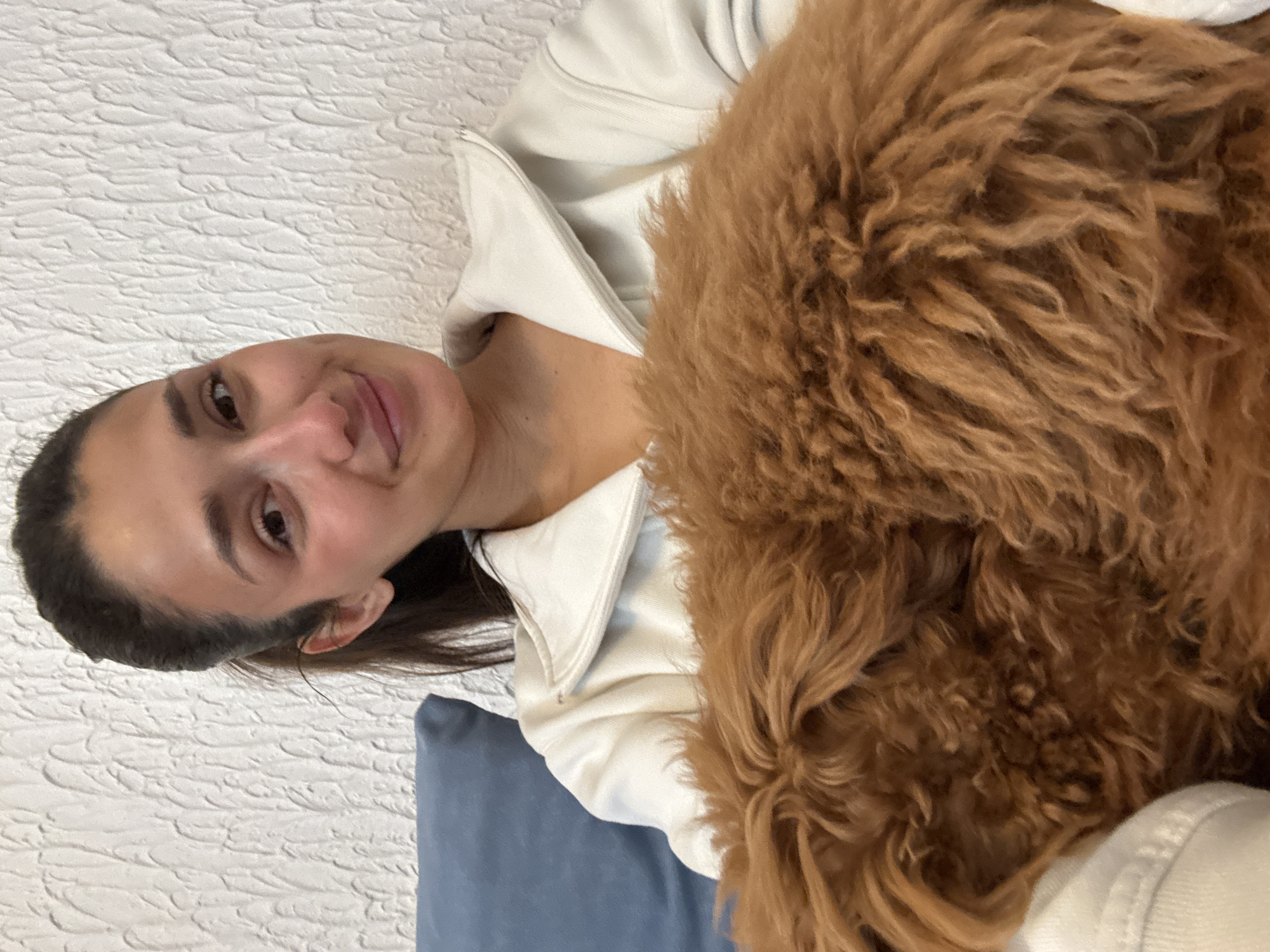 A woman with dark hair pulled back and wearing a white zippered jacket smiles at the camera. A brown, curly-haired dog is close to her, obscuring part of her jacket. They are on a blue pillow, resting against a textured white wall. The image could be taken indoors. The location of Sutomore, Montenegro, is not evident in the image.