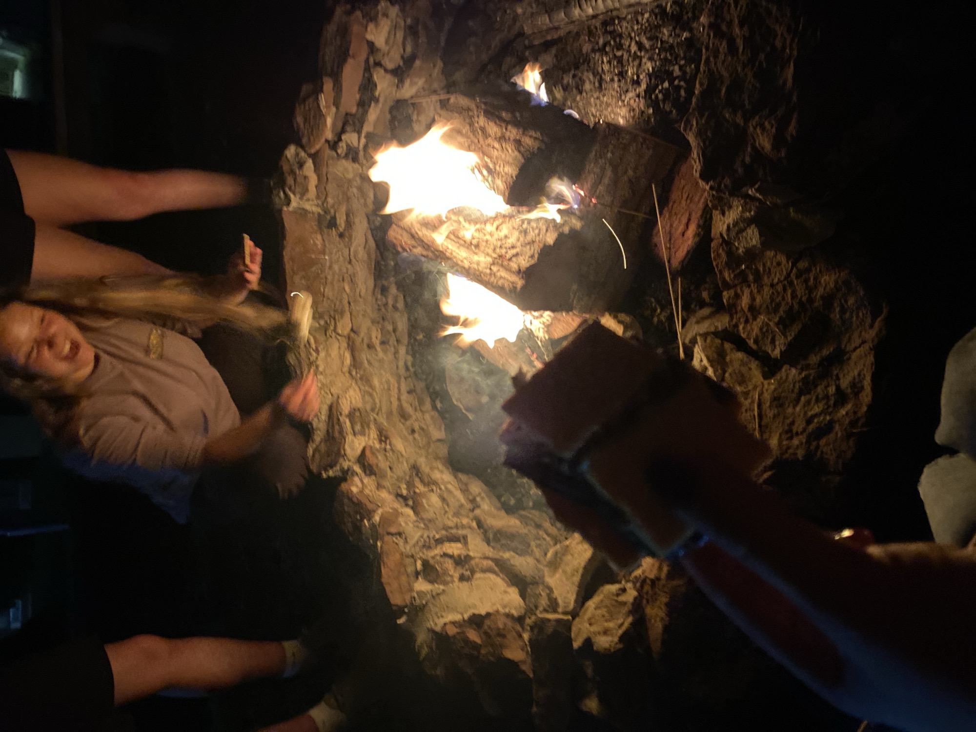 A low-angle, medium shot captures a campfire with burning logs at its center. The fire casts a warm glow on the surrounding rocks and smoke. In the foreground, a hand holds a skewered food item, possibly a s'more, towards the flames. A person's face, with an open mouth, is visible in the upper portion of the frame, suggesting they are reacting to the heat or participating in the activity. The legs of two individuals are also visible in the background. The scene appears to be an outdoor setting, possibly a backyard or campground, in Washington Township, United States of America.