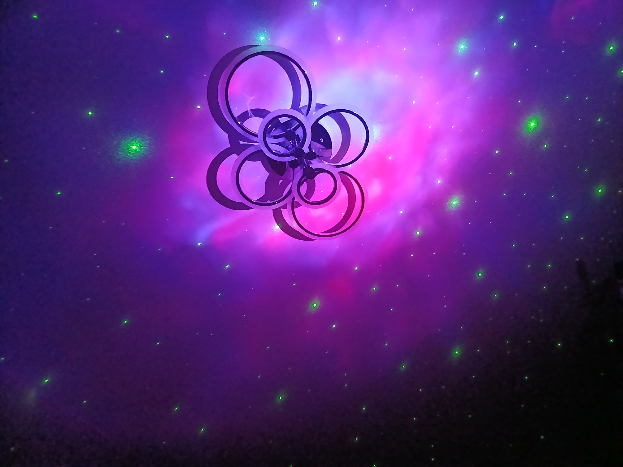 An abstract, abstract light projection displays a cluster of intersecting circles against a nebulous background of purple and pink hues, punctuated by scattered green specks that resemble stars. The circles are rendered in a dark, metallic material, creating a stark contrast with the vibrant, ethereal backdrop. The overall impression is one of cosmic wonder and artistic abstraction, evoking a sense of distant galaxies and celestial phenomena. There is no specific identifiable location in Chaikovsky, Russia, visible in this image; it appears to be a digitally rendered or projected scene.