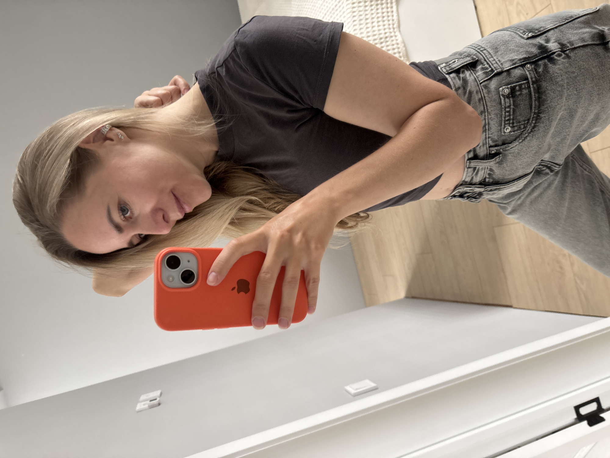 A woman with blonde hair is taking a mirror selfie in a room. She is wearing a dark grey crop top and light grey jeans. She is holding an orange iPhone in her right hand, which is angled towards the mirror. Her left arm is bent and her hand is resting on the back of her head. The background shows a white wall, a door, a wooden floor, and a bed with a textured white throw. The image does not contain any information about the city of Chaikovsky, Russia.