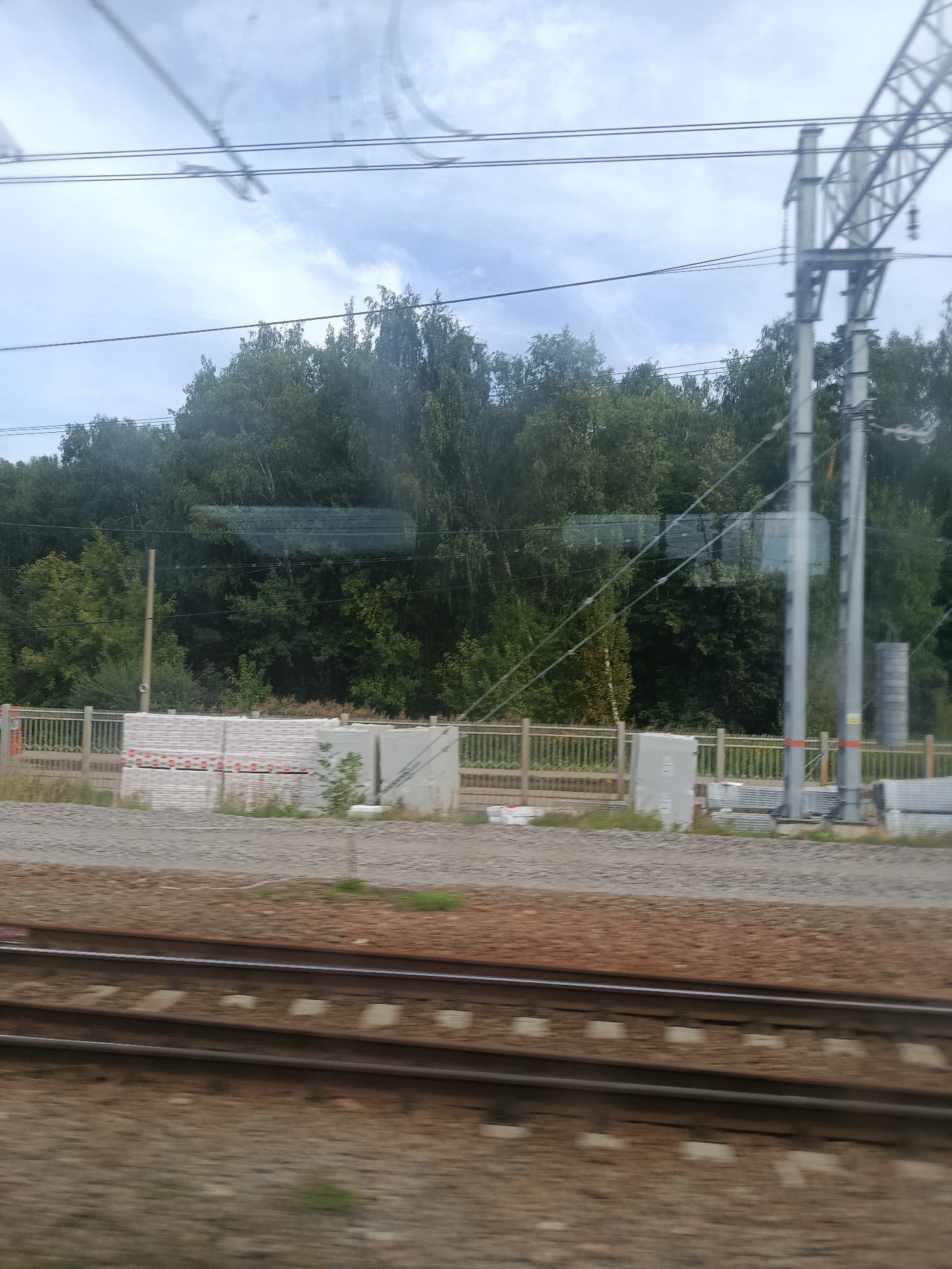 The image depicts a view from a moving vehicle, likely a train, looking out at a landscape that includes railway tracks in the foreground. A gravel bed with interspersed concrete sleepers supports the two parallel railway tracks. Beyond the tracks, a metal fence runs horizontally across the image, separating the railway area from a patch of land with stacked building materials, appearing to be packages of insulation or similar products, and some concrete structures. Further in the background, a dense line of deciduous trees forms a natural barrier, with tall, slender poles supporting overhead electrical wires for the railway, and a large metal pylon structure to the right. The sky above is overcast with light clouds, suggesting a temperate climate. The presence of railway infrastructure, including tracks and overhead lines, strongly indicates this scene is part of the Russian railway network. Given the specific mention of Balashikha, Russia, this view is likely captured along a railway line within or near this city.