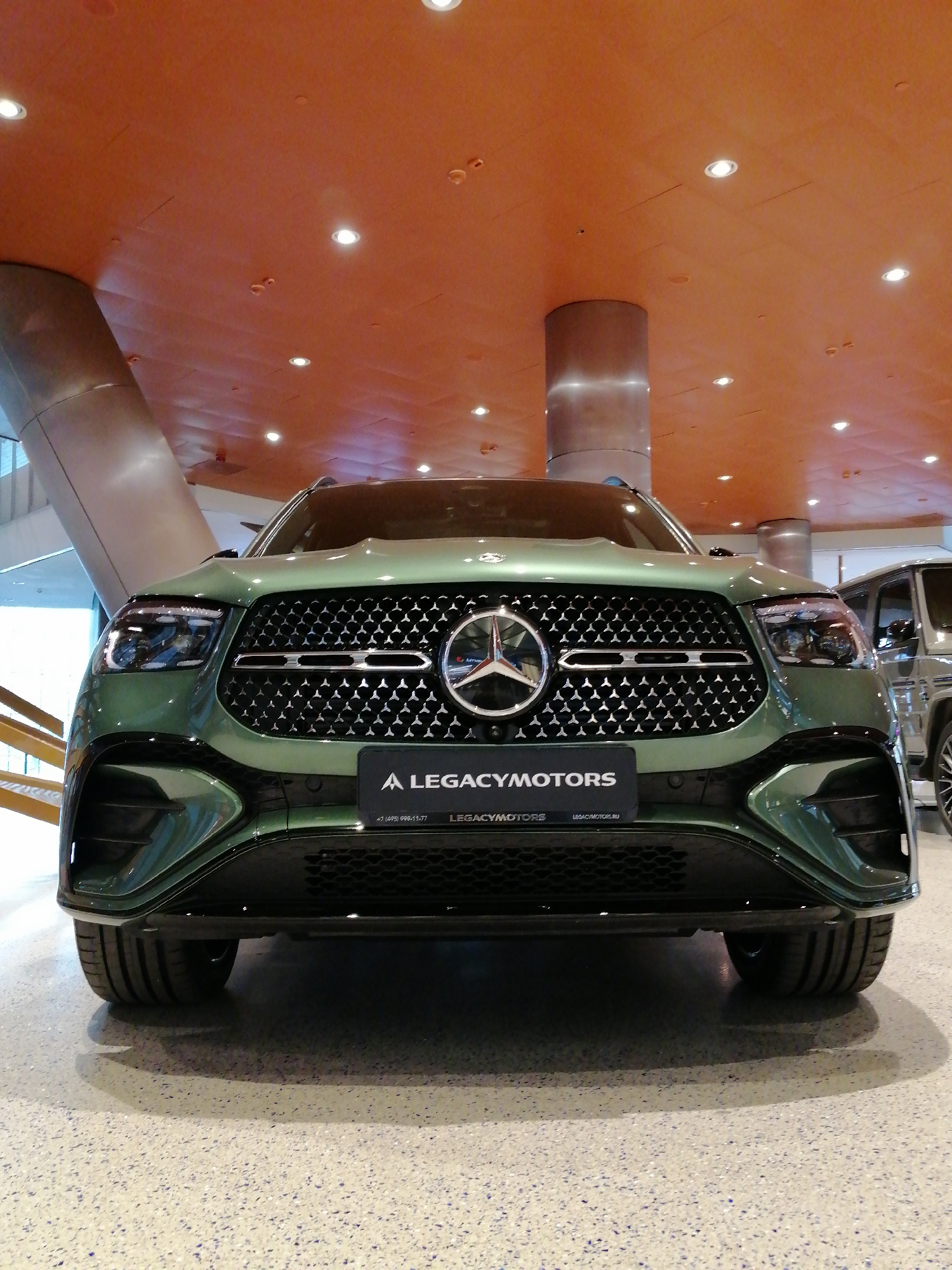 The image depicts the front of a green Mercedes-Benz GLE SUV, showcasing its grille and headlights. A dealership sign "A LEGACYMOTORS" is attached below the Mercedes emblem on the grille. In the background, parts of the dealership interior are visible, including structural columns and an orange ceiling with recessed lighting. Another vehicle is partially visible on the right side of the frame. The location is likely a car dealership in Moscow, Russia, given the sign's URL and the overall aesthetic. The ground appears to be a speckled gray floor.