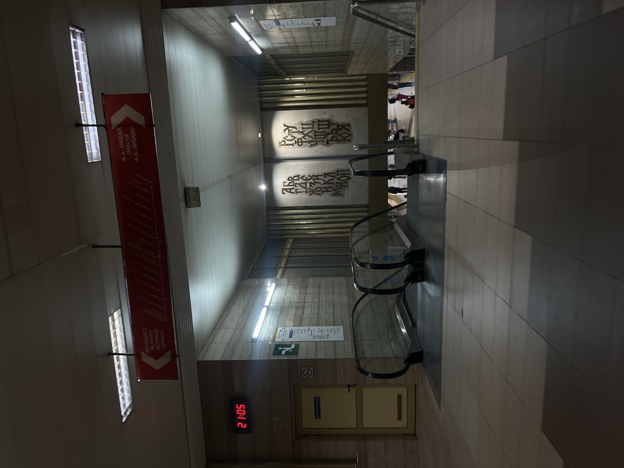 The image shows a subway station in Sofia, Bulgaria. It is a downward view of an escalator, flanked by tiled walls. At the top of the escalator, a landing shows people moving about. On the left is a red sign displaying station names. The walls are decorated with stylized lettering in Cyrillic. A digital clock displays the time 21:05.