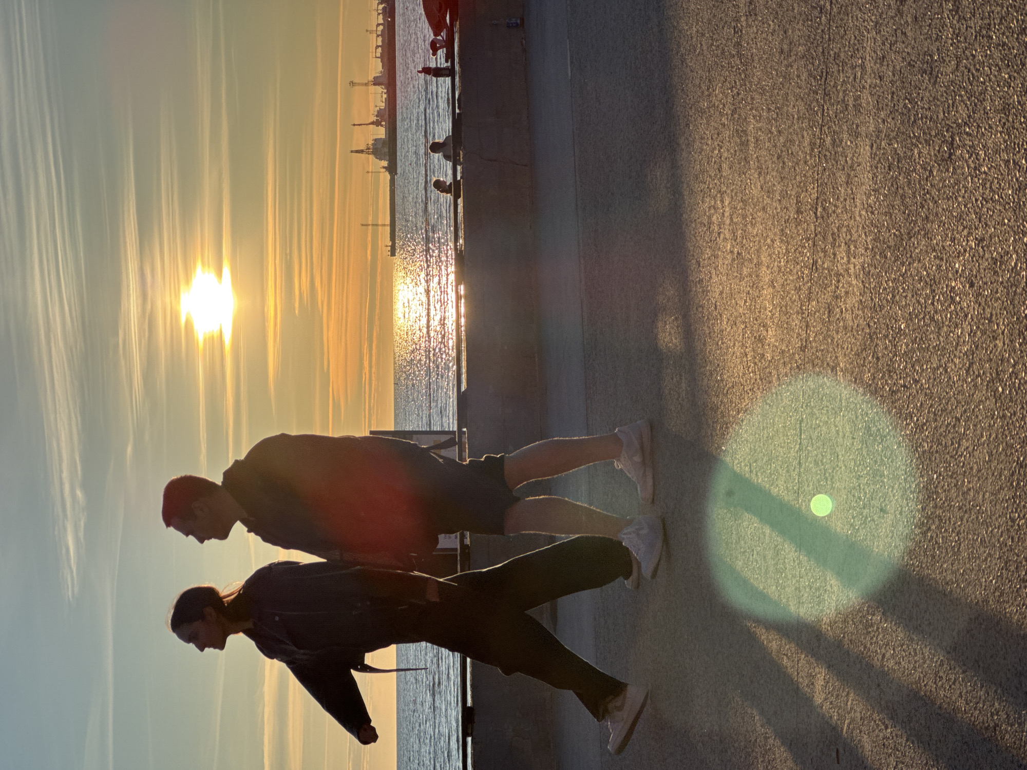 The image depicts two individuals walking along a promenade by the sea at sunset. The sun, a bright orb with visible rays, is low in the sky, casting long shadows and creating a golden sheen on the water and the paved ground. The sea is calm, with faint ripples reflecting the sunset's colors. In the background, a harbor with several cranes and industrial structures is visible on the horizon. A concrete barrier separates the promenade from the sea. The ground is a textured asphalt or similar material, showing a distinct line running across it. A circular lens flare, tinged with green and containing a bright central point, is present in the foreground on the ground. The overall scene conveys a sense of a peaceful evening stroll. This location appears to be the Tallinn Bay promenade in Tallinn, Estonia, given the characteristic industrial harbor silhouette visible.