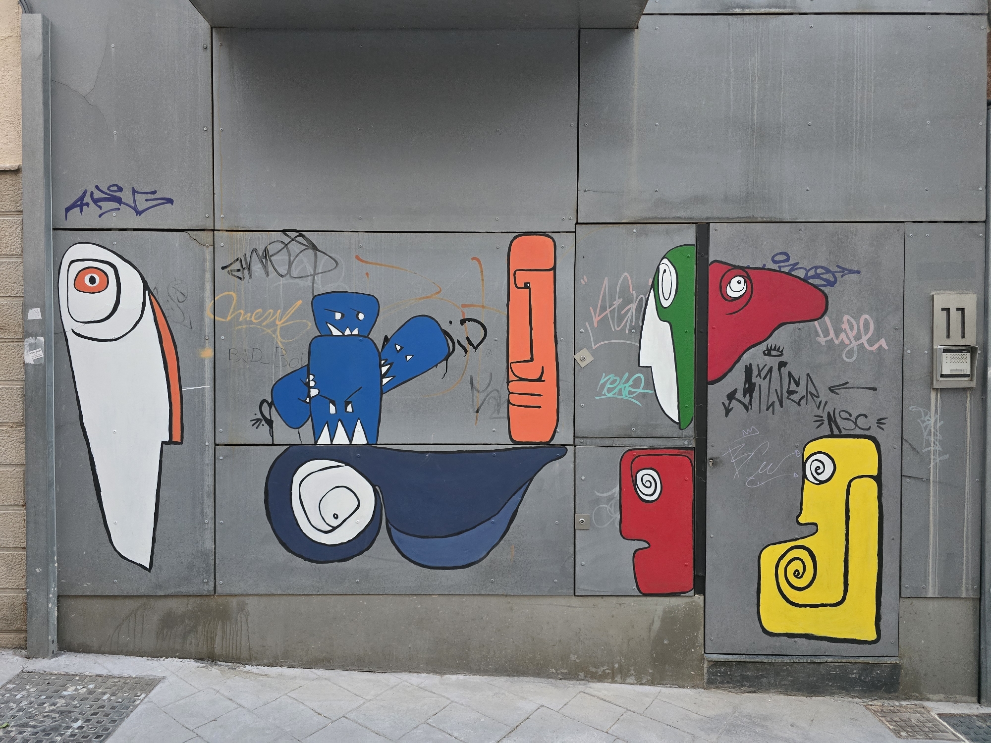 The image shows a section of a grey, metal-clad wall covered in various graffiti and street art. The artwork features several stylized, cartoonish figures. On the left, a large white figure with an orange stripe and a single red eye is prominent. To its right, a blue robot-like figure with sharp teeth is depicted. Below these figures is a large, dark blue shape with a white and black eye. Further to the right, an orange vertical figure with a black outline and facial features is visible. Adjacent to this, a figure with a split head, one side green and white, the other red, is painted. To the far right, a red rectangular figure with a white spiral eye and a yellow figure with a similar spiral eye are present. The wall is also marked with various tags and scribbles in different colors. A metal conduit runs vertically along the left edge of the wall. The lower section of the wall is a concrete base, and in front of it is a paved sidewalk with a metal grate. A building facade with light-colored brickwork is visible to the far left. The scene is illuminated by natural light. This artwork is located on the exterior wall of the former Mercado de Abastos in Jaén, Spain, now repurposed as a cultural space.
