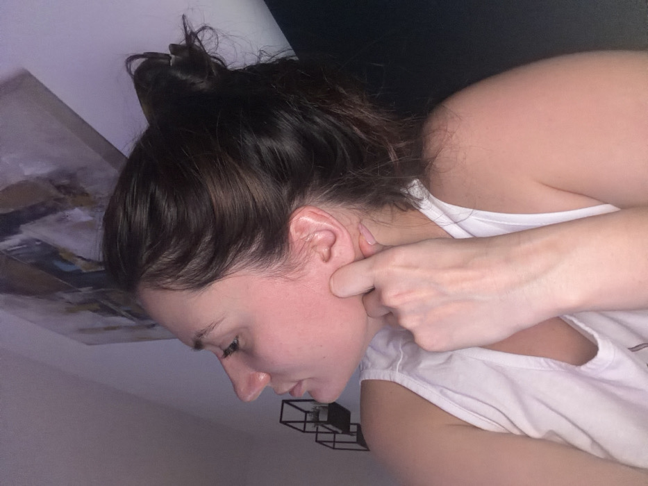 A woman in a white top is pictured from the side. Her brown hair is pulled back in a messy bun. Her face is angled downwards, and her eyes are closed. A hand is positioned on her face with a finger pressing against her cheek near her ear. An abstract painting is partially visible in the background on the left, and a metal geometric object appears near the bottom of the frame. The overall lighting is soft and slightly dim.