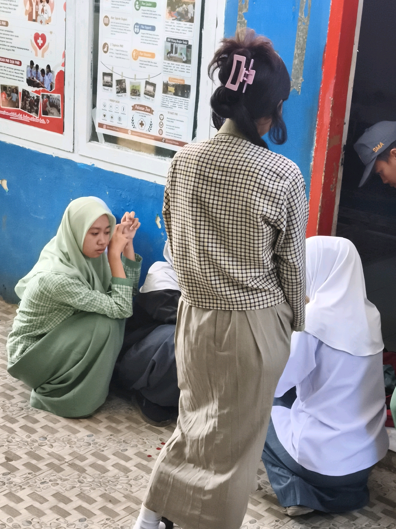 In Makassar, Indonesia, a young woman wearing a light green hijab and checkered shirt kneels on a patterned tiled floor. Her hands are clasped in front of her. To her right, another young person in a white hijab and light blue shirt is also kneeling. A third individual, seen from the back, wears a checkered shirt and a light brown skirt. They have dark hair styled with a pink clip. Behind them, a wall is painted in sections of blue, red, and black. A person wearing a gray baseball cap with "SMA" printed on it is partially visible. Posters are displayed on a white surface to the left. The scene appears to be outdoors or in a covered outdoor area.