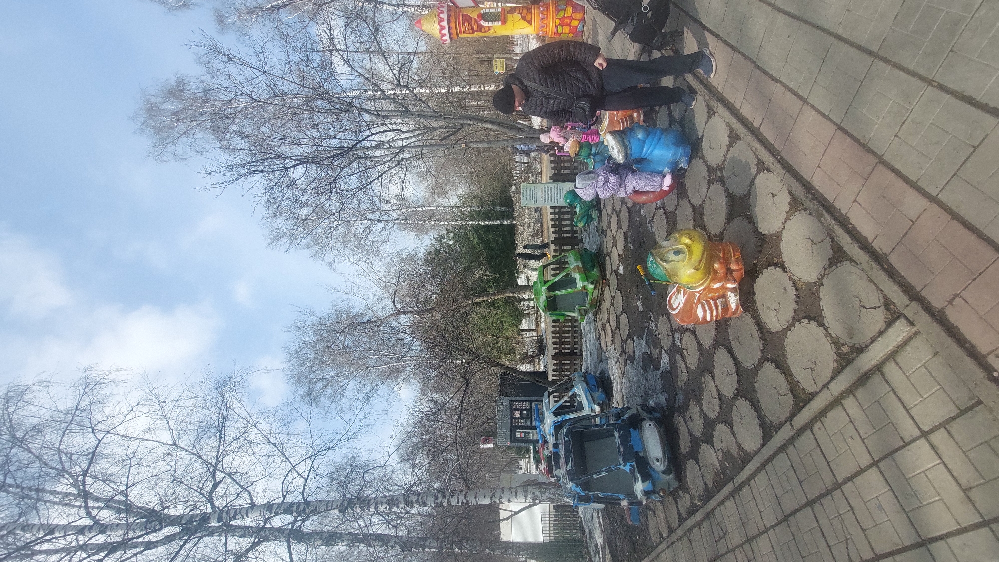 A ground-level shot shows an amusement park ride, a paved path, and leafless trees under a partly cloudy sky. A man in a dark jacket and pants stands near the colorful ride, which consists of multiple vehicles, including blue cars, green military-style vehicles, and animal-shaped seats. The ride is situated on a concrete surface patterned with circles resembling cross-sections of tree trunks. To the right, a paved path composed of rectangular paving stones borders the ride. Leafless trees dominate the background, extending towards the sky, which is partly obscured by clouds.
The location is identified as Novosibirsk, Russia.