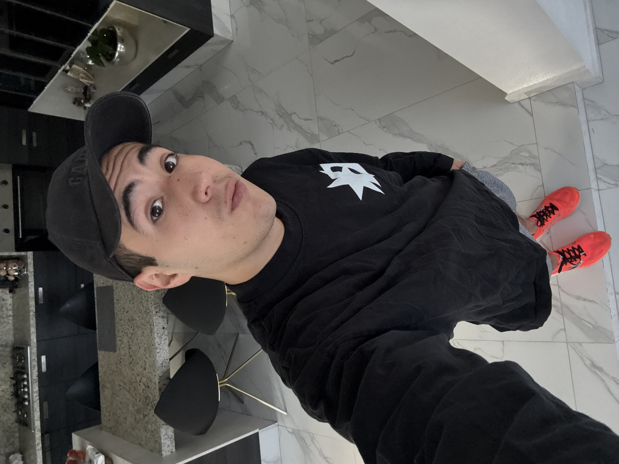 A young adult male, likely in his late teens or early twenties, stands upright in what appears to be a modern kitchen or open-plan living area located in San Bernardino Tlaxcalancingo, Mexico. He is positioned centrally, looking directly at the camera with a neutral expression, and is taking a selfie. He wears a dark grey baseball cap with "GAP" branding, a long-sleeved black shirt featuring a white stylized logo on the left chest, grey patterned shorts, and bright neon orange athletic shoes with black laces over grey socks. His right hand holds the camera, and his left hand is tucked into his pocket.
The flooring consists of light-colored, possibly marble or marble-look, tiles with grey veining. In the background to the left, a kitchen island or counter with a speckled light grey granite or quartz surface is visible, under which two black bar stools with gold-colored metal bases are placed. Behind the counter, there are dark grey or black kitchen cabinets and drawers, a stovetop with multiple burners, and control knobs. To the right, a dark, reflective appliance (possibly an oven or microwave) is integrated into the cabinetry. Further right, a white and black open shelving unit displays decorative items, including a silver-colored bowl containing a green plant. A plain white wall or column borders the right side of the frame. The lighting is uniform, suggesting interior artificial illumination.