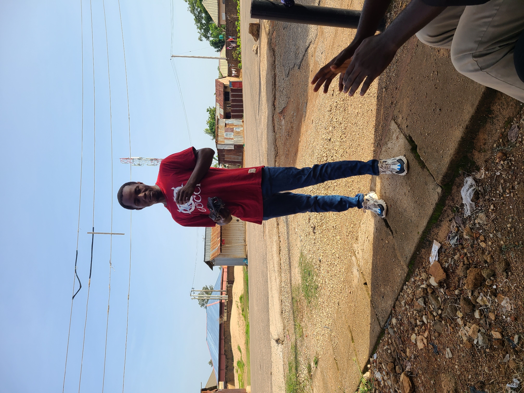 An individual stands outdoors in what appears to be an urban or semi-urban setting in Gura Top, Nigeria. The person is wearing a red t-shirt with a white graphic and blue jeans, and is holding a dark object, possibly a camera or phone, in their hands. Another person's hands are visible in the foreground on the right, appearing to clap. The background features a road, scattered buildings with corrugated metal roofs, and utility poles with overhead wires. The ground is a mix of dirt, gravel, and some debris. The sky is clear and blue.