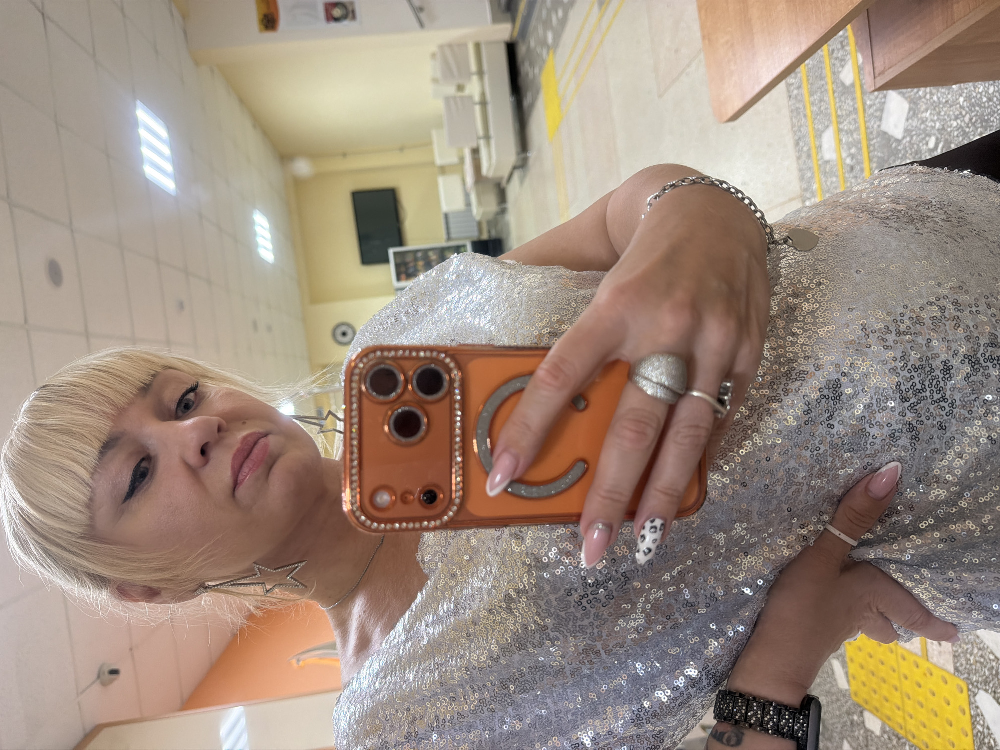 The image shows a blonde woman with bangs taking a selfie in what appears to be an indoor public space. The woman is wearing a sequined silver top or dress and large star-shaped earrings. She is holding an orange phone with a bejeweled case to take the picture. Her fingernails are painted with a variety of patterns. She has rings on several fingers and a watch on her left wrist. The background includes rows of chairs and yellow stripes on the floor, suggesting a waiting area. In the distance, there is a television screen and what looks like vending machines. The location is in the city of Chaikovsky, Russia.