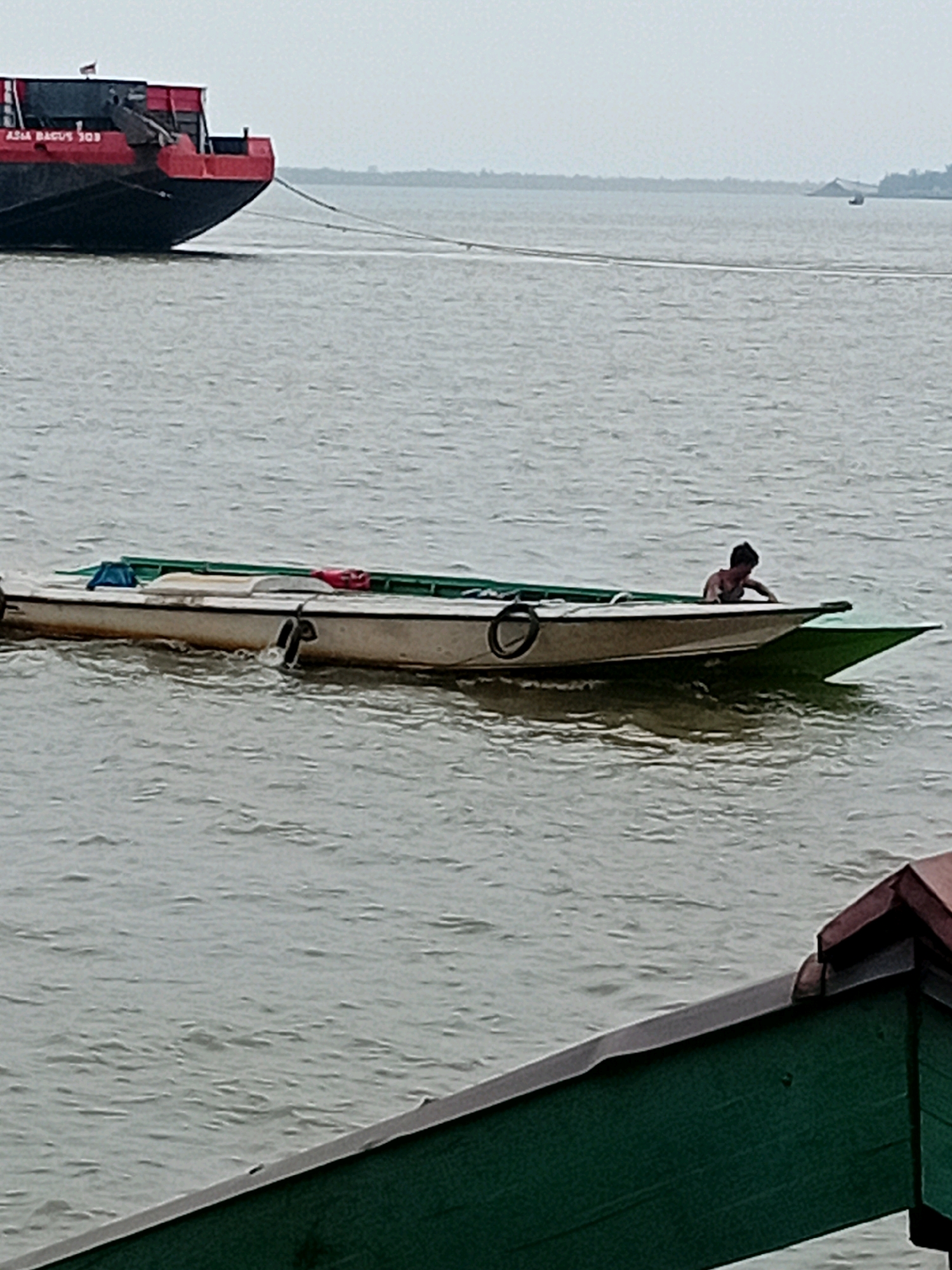 A large barge, painted red and black with "ASIA BAGUS 303" written on its side, is partially visible on the left side of the frame. It is tethered by a rope to a smaller boat in the center of the image. The boat is a long, narrow vessel, light-colored with green trim, and appears to be partially submerged. A person, with dark hair and wearing a dark top, is leaning over the side of the boat, appearing to be tending to something or preparing to dive. The water is calm and grey, with ripples indicating a gentle movement. In the background, a hazy shoreline with trees is visible, suggesting the location is a river or coastal area. The foreground shows a portion of a green structure with a dark trim and a reddish-brown roof, possibly a dock or part of a building. The sky is overcast and light grey. The scene depicts a man in a boat connected to a larger vessel in a waterway.