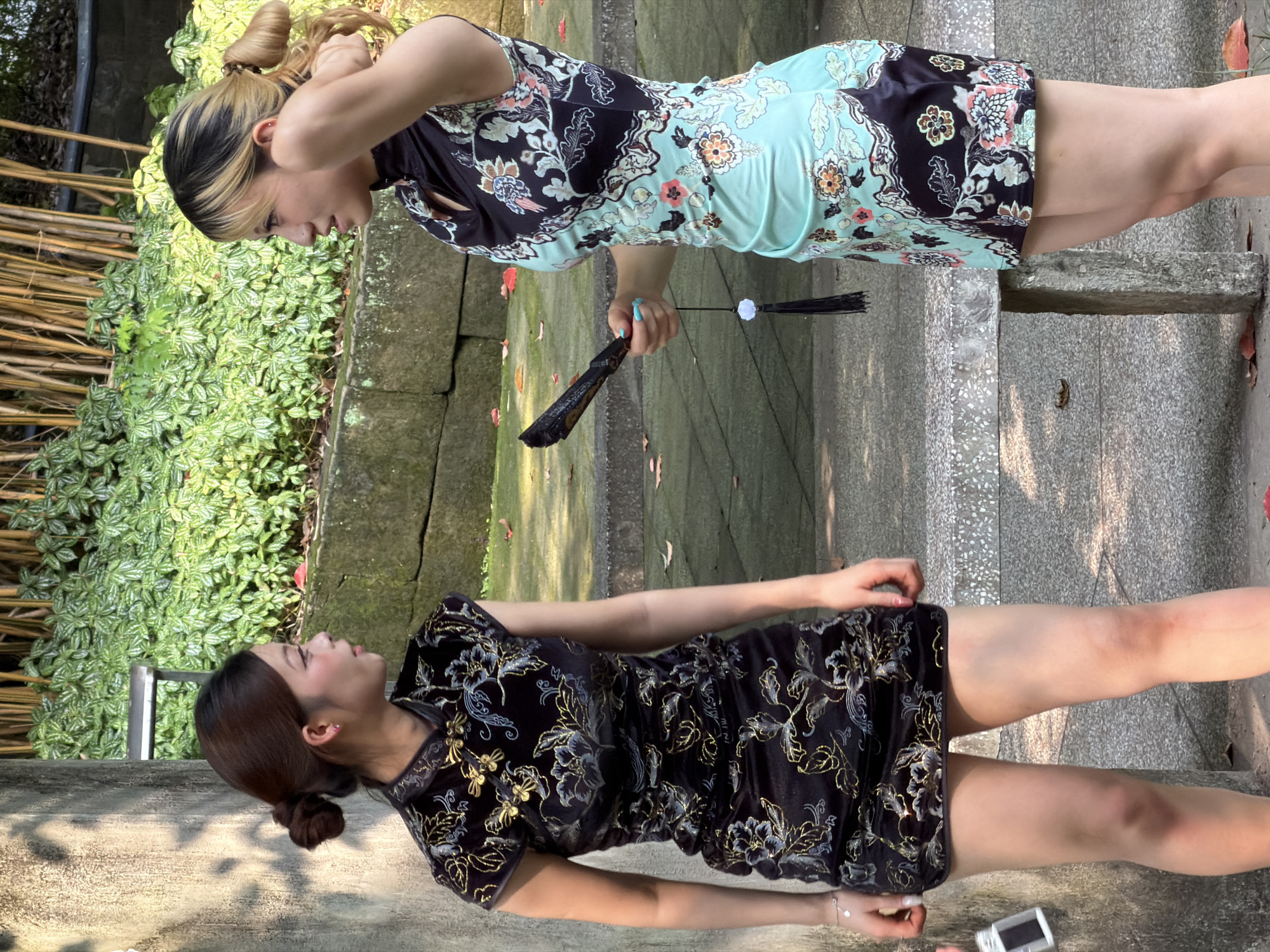 Two East Asian women are observed standing and interacting in an outdoor setting, understood to be in Yuzhong District, China. The woman on the left, with dark hair styled in a bun, wears a short-sleeved, high-collared, form-fitting black dress with gold and blue floral patterns. A small silver object is partially visible in her left hand. She faces right, looking towards the second woman. The woman on the right, with blonde-streaked hair pulled back, wears a sleeveless, high-collared, form-fitting dress that features a black upper section with floral and avian patterns and a light teal lower section with similar floral designs. She holds a black folding fan with a black tassel and white bead in her right hand, while her left hand is raised to the back of her head. She faces left, looking towards the first woman. Both individuals appear engaged in a direct conversation. The immediate background includes a light grey concrete pillar to the far left, a low stone wall with dense green foliage behind both women, and multiple slender bamboo stalks further behind the foliage. The ground beneath them is paved and sparsely covered with fallen reddish-brown leaves. The scene is illuminated by bright natural sunlight, creating distinct shadows.