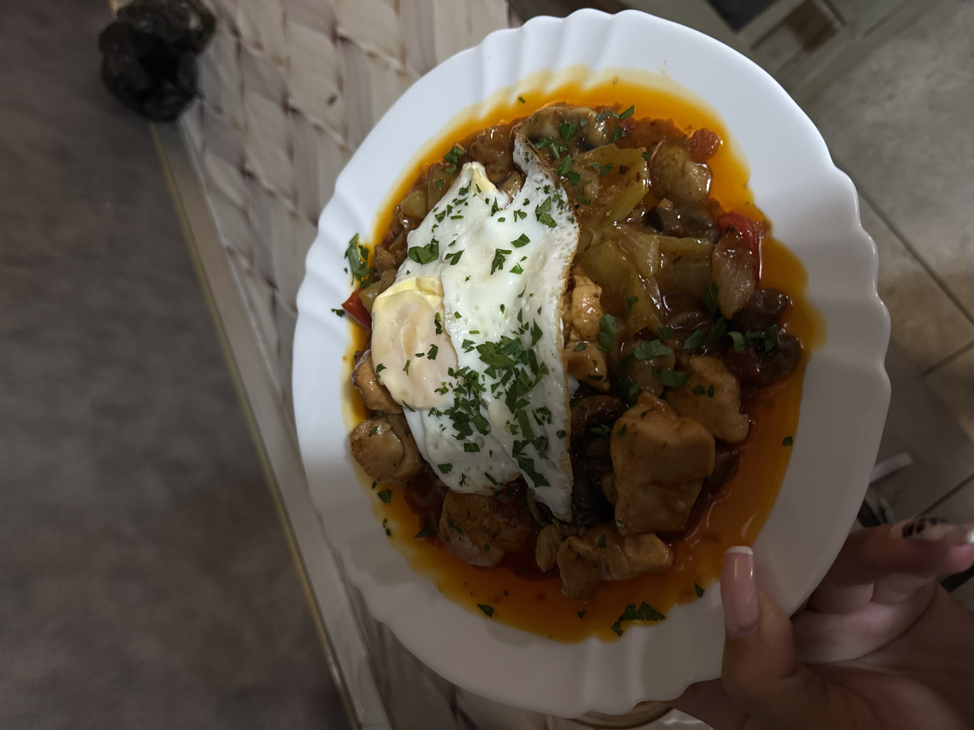 The image shows a close-up, top-down view of a white plate filled with a meal. The meal consists of cubed chicken, vegetables such as peppers and mushrooms, and a fried egg served on top. Chopped parsley is sprinkled over the dish. The plate has a decorative, wavy edge.
The plate is placed on a surface that appears to be a tiled countertop, with a metallic edge visible. In the background, a dark, lumpy object is visible on the left side, possibly trash or a bag. The lighting suggests an indoor setting, possibly a kitchen or dining area. A person's hand with manicured nails is holding the edge of the plate on the right side of the image. There is no visible text in the image. The overall impression is of a home-cooked meal, ready to be eaten.