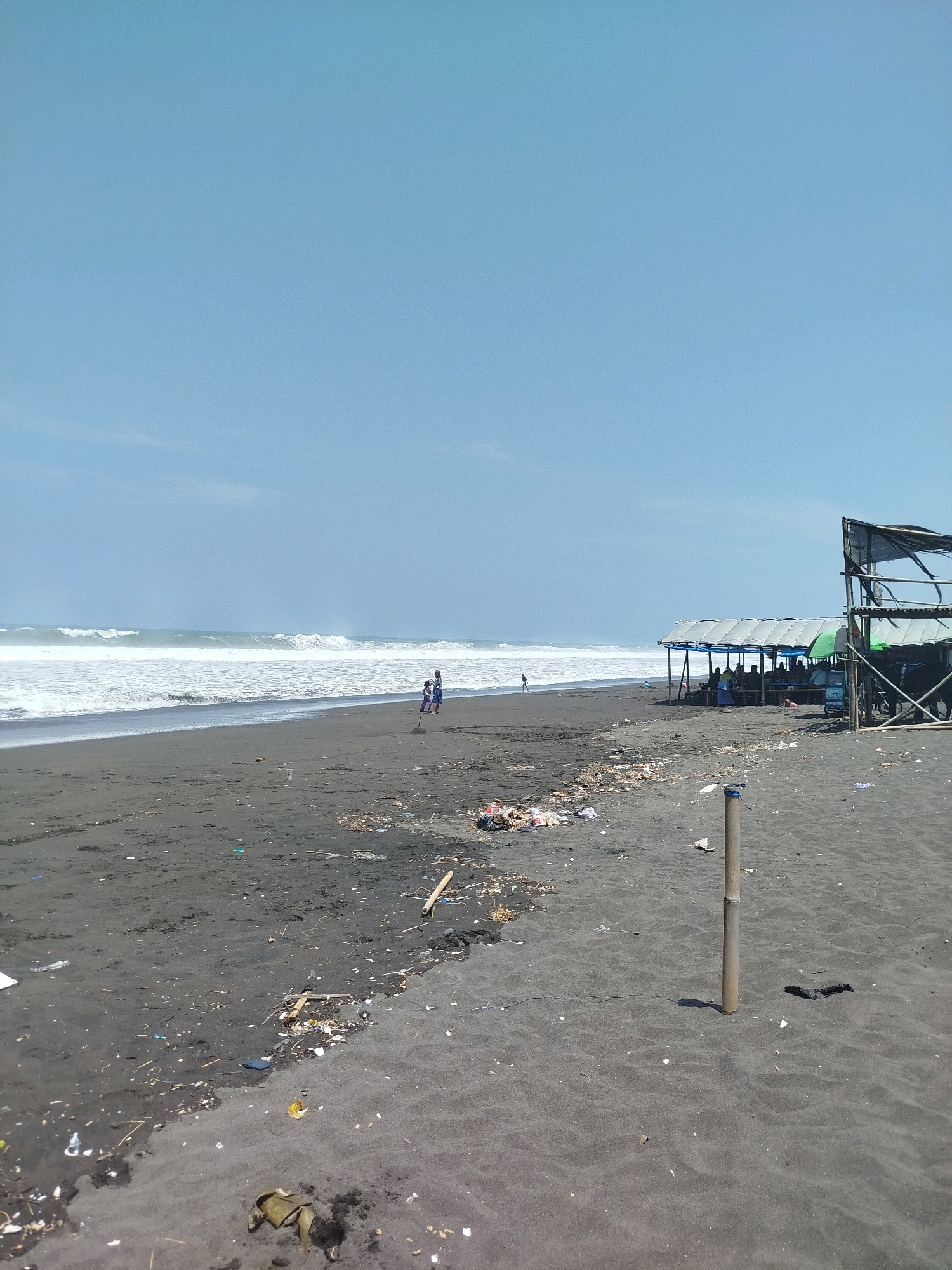 This is a photograph of a beach in Buluspesantren, Indonesia. The beach has dark sand and is littered with various types of trash, including plastic bottles, wrappers, and wooden debris. In the background, the ocean has large waves crashing onto the shore under a clear blue sky. There is a simple open-air shelter with a metal roof and a few people sitting underneath it, suggesting it might be a small vendor or seating area for beachgoers. To the right of the shelter, there is a more permanent structure with a corrugated metal roof supported by wooden poles. A single bamboo pole stands upright in the sand in the foreground, casting a shadow. Two figures, possibly children, are visible on the beach near the water's edge, one with a broom. Another lone figure can be seen walking further down the beach.