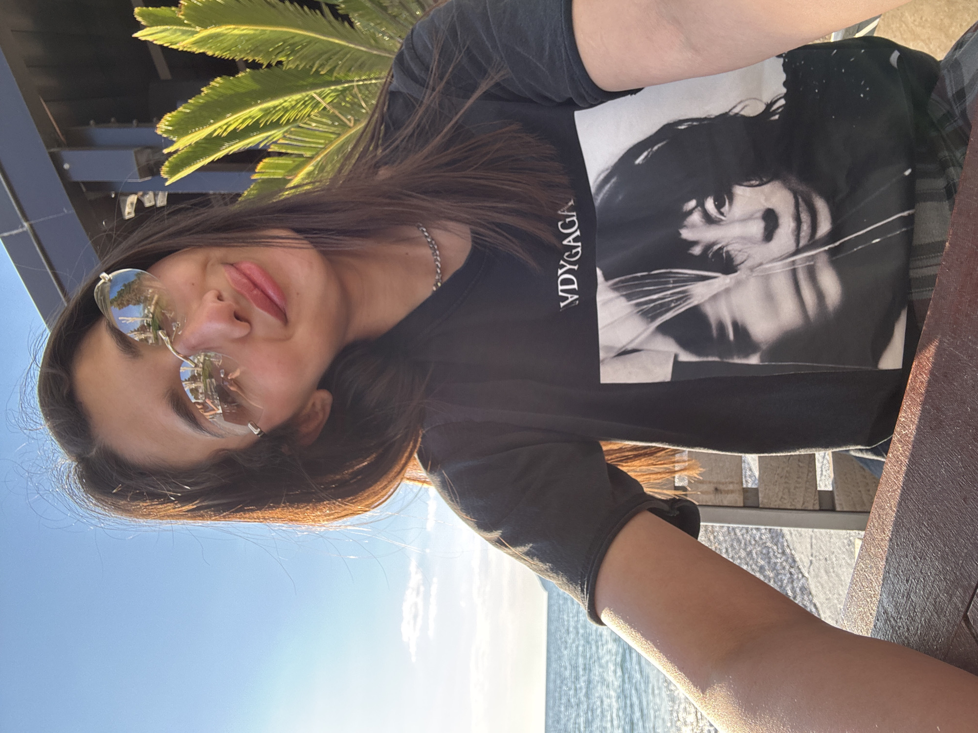 A young woman is depicted in a selfie-style photograph, taken outdoors under a clear, bright blue sky with sparse white clouds. She has long, dark hair, wears reflective, round sunglasses, and a thin silver chain necklace. Her lips are a soft pink, and she has a subtle smile with her tongue slightly visible. She is wearing a black t-shirt that features a prominent black and white portrait of a person, with the text "VDY GAGA" printed vertically on the side. Her arm, visible in the foreground, rests on a dark wooden surface, likely a table or railing, which also shows part of a plaid pattern, possibly from shorts or pants.
In the background, lush green palm fronds are visible in the upper left, hinting at a warm, coastal environment. Beyond the wooden surface and the woman's arm, a calm body of water, presumably the sea, extends towards the horizon. The bright sunlight casts a warm, golden glow on her skin and the wooden surface, suggesting it is daytime, possibly late afternoon or early morning. The overall scene conveys a relaxed atmosphere in Bar, Montenegro.