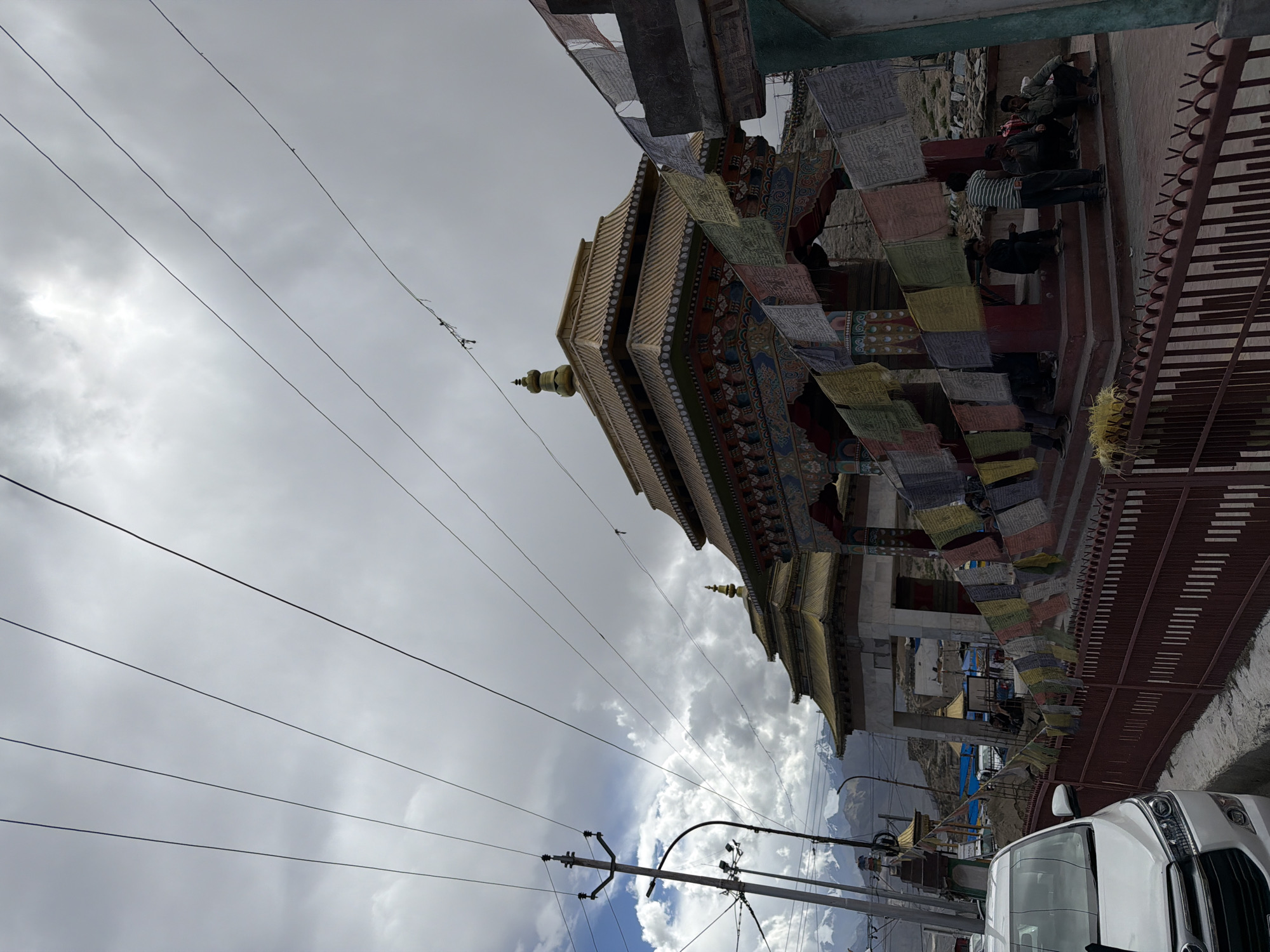 The image depicts the entrance to a monastery in Leh, India, under a cloudy sky. The architecture features ornate, colorful detailing and golden roofs, characteristic of Himalayan Buddhist monasteries. Prayer flags, in various colors and inscribed with text, are strung across the entrance and around the structures, fluttering in the wind. Several individuals are seated on the steps leading into the monastery, some appearing to be in conversation. In the foreground, a white car is partially visible, parked on the left side of the frame. Behind the monastery, a vast mountain range with snow-capped peaks is visible, indicating the high-altitude location. Electrical poles and overhead wires traverse the sky, suggesting the presence of infrastructure in the area. The overall scene conveys a sense of cultural and spiritual significance, set against a dramatic natural landscape.