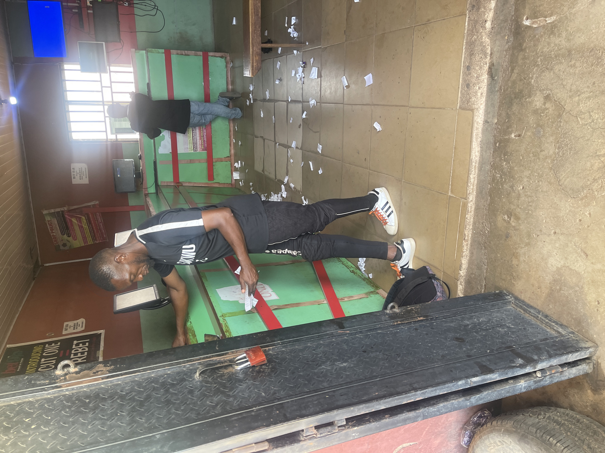 The image depicts the interior of a betting shop in Gura Top, Nigeria. A man in black sportswear is near a counter, appearing to be sorting or checking slips of paper. Another man wearing a dark jacket and jeans is visible in the background, standing near a row of betting terminals. The floor is littered with discarded paper slips. Several betting terminals, some displaying illuminated screens, line the wall. Promotional posters are displayed on the wall behind the counter. The entryway to the shop features a heavy black metal door secured with a padlock. A tire is visible near the entrance. The general appearance of the interior suggests a modest, functional establishment.