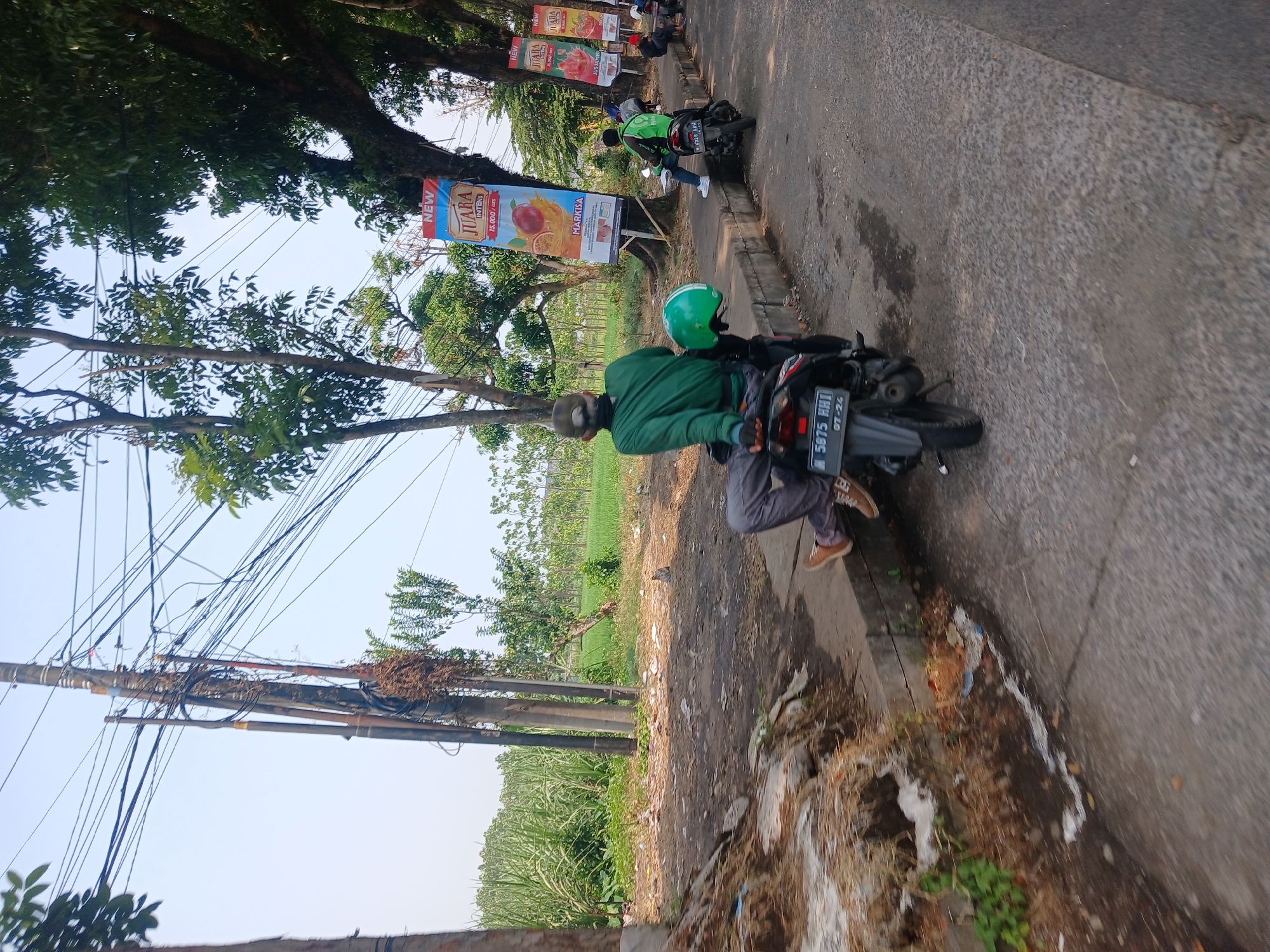 The image depicts a street scene in Singosari, Indonesia. In the foreground, a person wearing a green long-sleeved shirt and grey pants is sitting on a black motorcycle, facing away from the camera. The motorcycle has a license plate that reads "N 5875 HHI 07-24". A green helmet rests on the back of the motorcycle. The person is wearing brown shoes. Behind the motorcycle, another person in a green jacket is sitting on a similar motorcycle. Several banners are visible on the right side of the image, displaying text that appears to be in Indonesian, advertising products like "JUARA INTENSE" and "JUS SEMANGAT". The environment is characterized by a sidewalk, a paved road, and a grassy area with trees and power lines overhead. In the background, there are fields of tall green crops, likely sugarcane, and smaller trees. The sky is clear and bright, suggesting a sunny day. The overall impression is of a typical roadside scene in a semi-rural or suburban area of Indonesia.