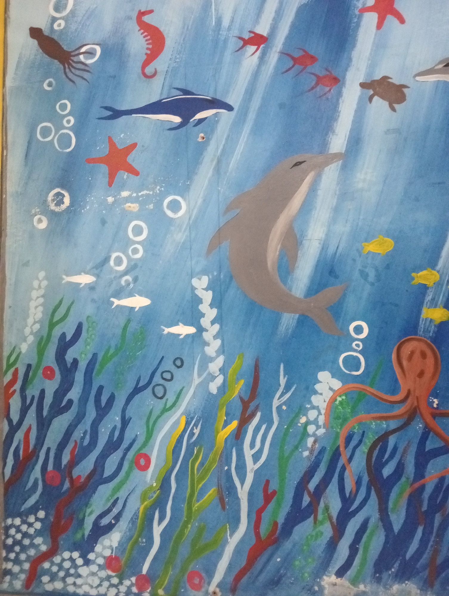 This is a colorful mural of an underwater scene, painted on a wall in Pringsewu, Indonesia. The mural depicts a variety of marine life swimming in blue water with stylized seaweed and bubbles. Visible creatures include a gray dolphin, a blue dolphin, a red seahorse, a brown turtle, an octopus with orange tentacles, a squid, and several small red and yellow fish. There are also white and red starfish. The background is a gradient of blue, suggesting the depth of the ocean, with white streaks that might represent light filtering through the water. The style is simplified and cartoonish, likely intended for a public space or educational setting. The mural appears to be in good condition, with vibrant colors.