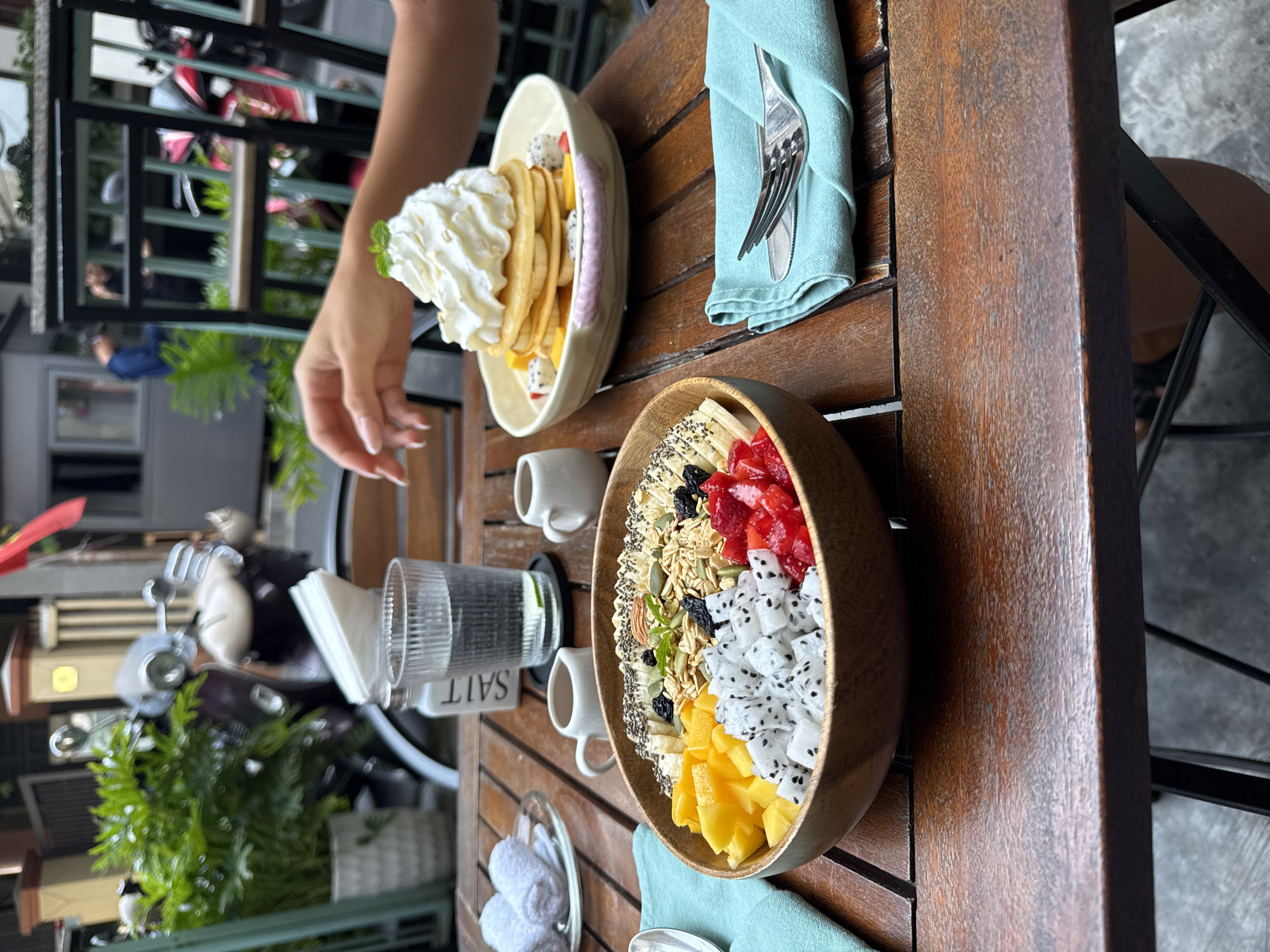 The image shows a wooden table laden with breakfast fare, likely in an outdoor cafe in Đà Nẵng, Vietnam. A person is reaching towards a plate of pancakes topped with whipped cream. A large wooden bowl holds a fruit salad with mango, dragon fruit, strawberries, bananas, and granola. Utensils are neatly placed on a light blue napkin. A glass of water and salt and pepper shakers are also on the table. The background shows a street with motorcycles and pedestrians, typical of Vietnamese cityscapes.