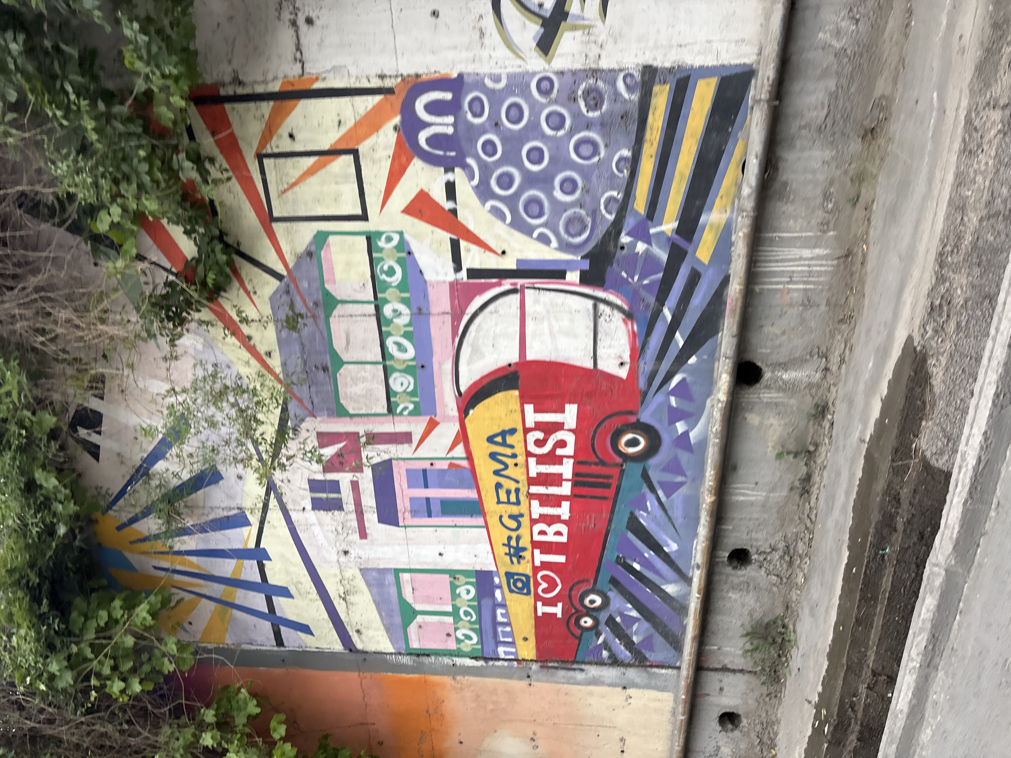 A wall mural depicting a stylized city scene is visible. The mural is painted on a concrete wall partially obscured by vegetation. The scene includes building facades rendered in pastel colors, a red bus with the phrase "I <3 TBILISI," and a pattern of purple circles. The hashtag "#GEMA" is also visible. The location of the image is Tbilisi, Georgia.