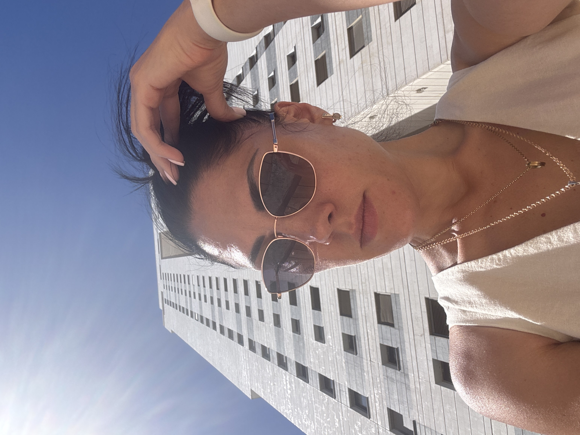 A woman wearing sunglasses and a white top is pictured in a close-up, low-angle shot with a high-rise building and blue sky in the background. She is touching her hair with her hand and wearing a wristband on her left wrist and a gold necklace. The building has a grid-like facade with repetitive window patterns, and the sun is shining brightly. Based on the architecture and background elements, this photo likely features the building at 17 Ha-Rav Kook street, Netanya, Israel.