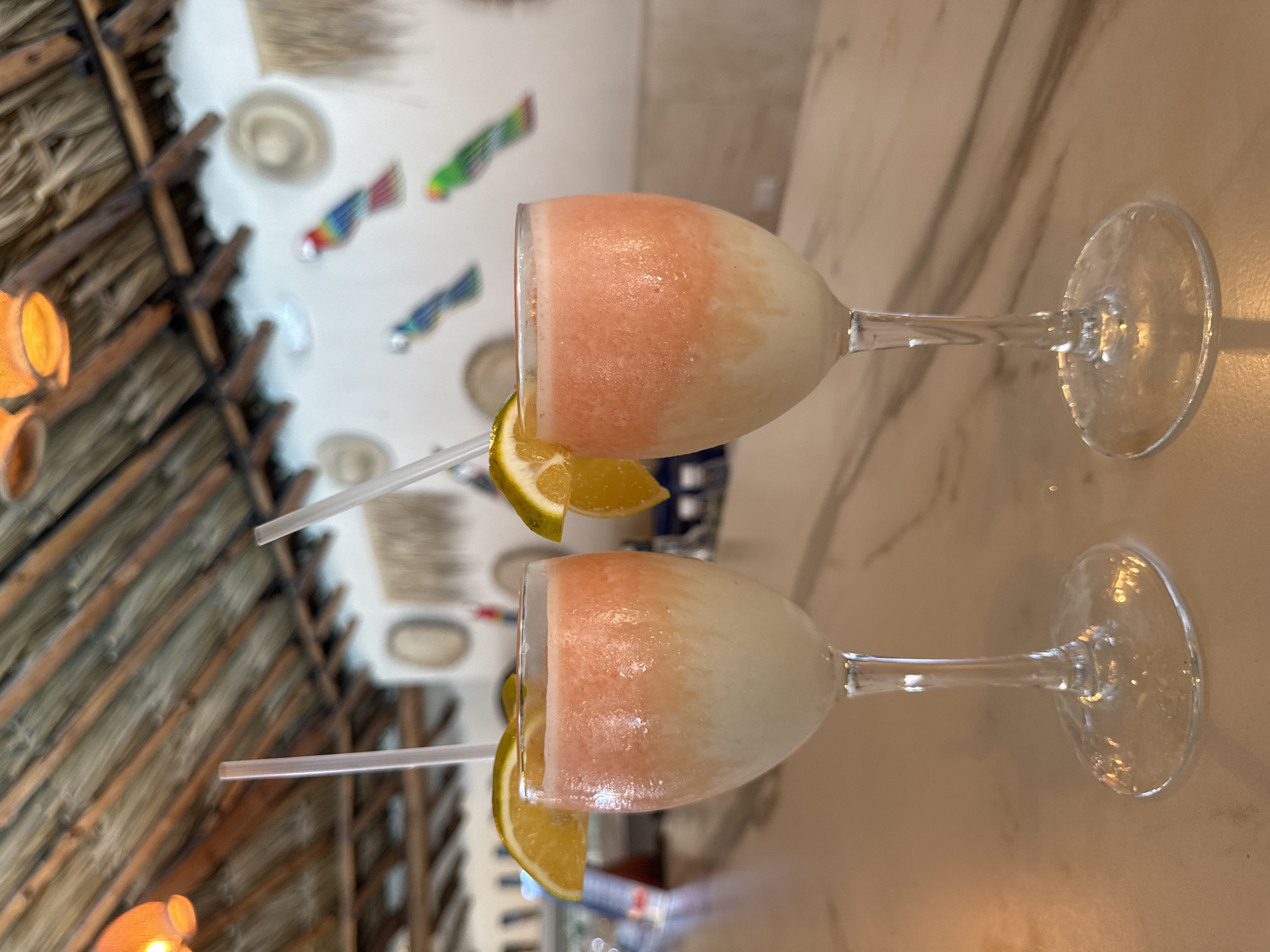 This image features two refreshing, two-toned frozen drinks presented in stemmed glasses, rotated 90 degrees clockwise. Each glass contains a beverage with a lighter white or pale yellow base and a vibrant pink or orange top layer, indicating a layered or blended frozen concoction. A slice of lime or lemon garnishes the rim of each glass, and a clear plastic straw is inserted into the drink. The glasses appear frosted, suggesting the drinks are very cold. They are placed on a smooth, light-colored surface, possibly marble or polished tile, which reflects some light.
In the blurred background, architectural elements suggest a tropical or rustic setting. A ceiling or roof structure made of interwoven thatch and wooden beams is visible on the left. Further back, a white wall is adorned with decorative elements, including several small, round, hat-like objects and a series of colorful, stylized figures that resemble fish or birds. The overall scene evokes a relaxed atmosphere, likely in a resort, restaurant, or bar in a tropical location, aligning with the provided context of El Tirano, Venezuela. The lighting appears to be indoor. No discernible text is visible in the image.