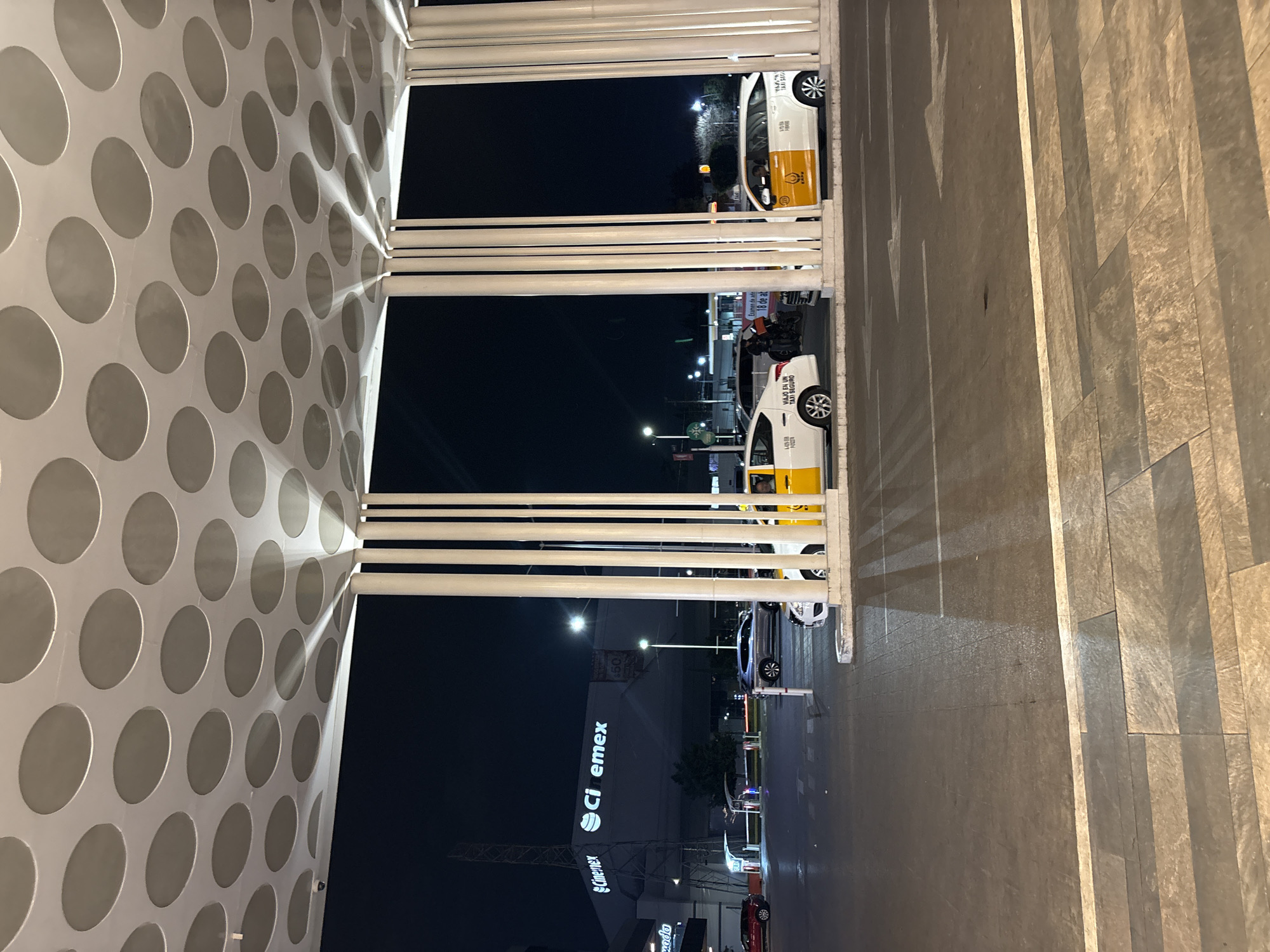 The image shows an exterior view, likely taken at night, capturing a scene outside a building with a facade featuring a pattern of repeating circles. The perspective is from an elevated position, looking down at street level. There are white horizontal bars visible in the foreground, possibly part of a railing or architectural structure.
On the street, there are several white and yellow taxis parked. A Cinemex sign is visible in the background, which helps identify the location as the city of Puebla, Mexico, as Cinemex is a prominent cinema chain in Mexico. Other signs and lights are visible in the distance, indicative of an urban environment at night. The road is paved, and there is a walkway or sidewalk on the right side of the frame, constructed of large, rectangular tiles. The lighting suggests that the photo was taken under artificial streetlights, and some of the surfaces reflect the light, indicating they may be wet or polished. One taxi visible has the text "VIAJO EN UN TAXI SEGURO" on its door.
