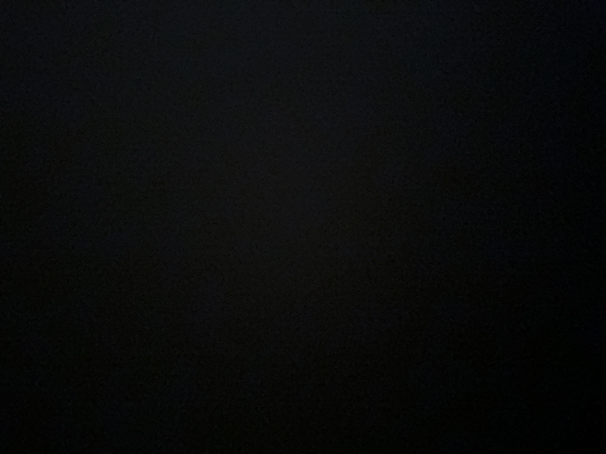 The image is a close-up, black and white photograph of a textured surface. The texture appears to be a rough, grainy material, possibly fabric or a painted wall. The lighting is very low, creating deep shadows and making it difficult to discern specific details. There are no discernible subjects, objects, or scenes. The image is entirely black with subtle variations in tone that suggest a textured surface. No activity or event is taking place. There are no visible text elements. The time of day, weather, location cues, and emotions are not discernible from the image.