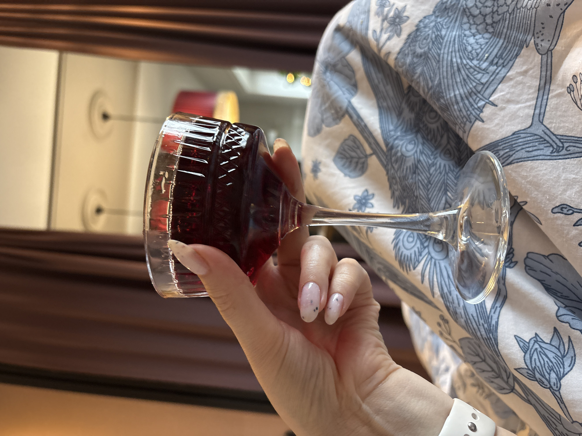 A hand, wearing a white smartwatch, holds a glass of red wine. The glass is short and wide with vertical ridges. The hand has manicured nails with a light pink base and small black specks. The backdrop is a light-colored fabric printed with blue-grey bird and floral designs. In the blurred background are hints of a room interior with brown curtains and a white wall with a reflection. The location is Saint Petersburg, Russia.