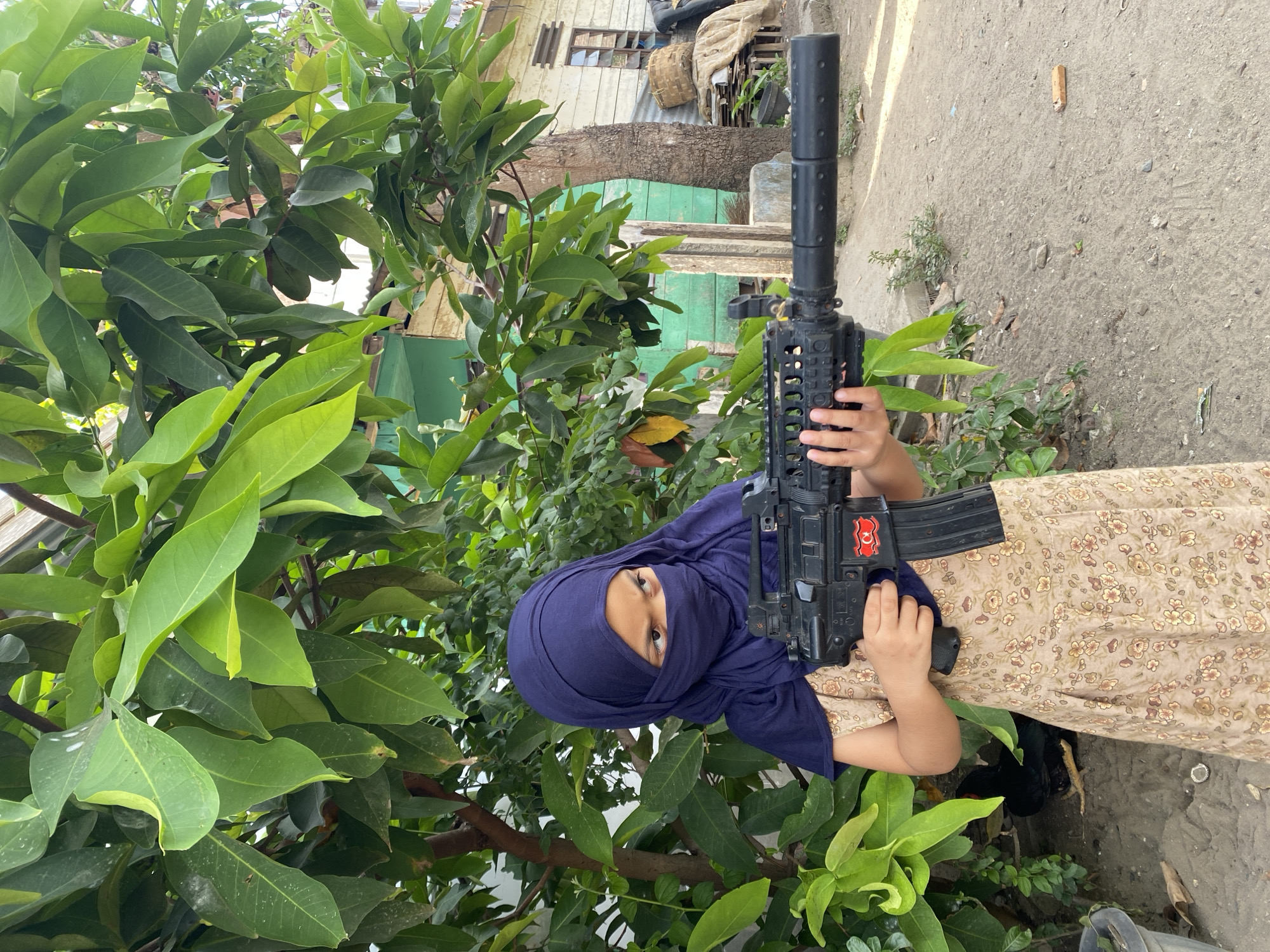 A child wearing a blue headscarf holds a black toy assault rifle in a cluttered outdoor environment. Lush green foliage surrounds the child, partially obscuring the view. In the background, a dilapidated structure with a wooden exterior and green accents is visible. The ground is concrete, showing signs of wear and debris. The photograph appears to have been taken at an angle and is presented in a rotated orientation. The location is presumed to be the city of Lhokseumawe, Indonesia.