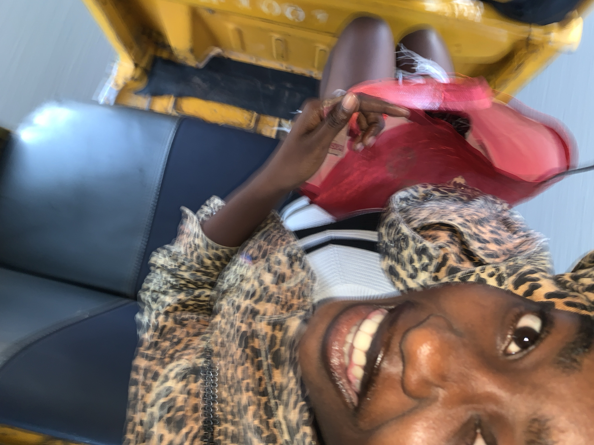 The image shows a close-up, vertically oriented, blurred selfie taken inside what appears to be a vehicle. A person with dark skin, wearing a leopard print top, is prominently featured, with their face tilted towards the camera and their mouth open in a smile, revealing teeth. Their right hand, holding a bright red bag, is visible in the foreground, reaching towards the bottom of the frame. The background shows dark blue seating and yellow interior panels of the vehicle, possibly a type of local taxi or bus, common in cities like Gura Top, Nigeria. The overall lighting is bright, suggesting daylight. The focus is sharpest on the individual's face and hand, while the surroundings are out of focus due to the motion blur.