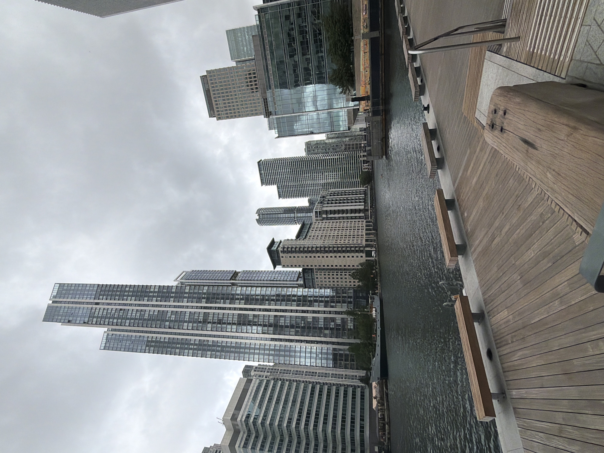 The image, correctly oriented, captures a modern urban waterfront scene under an overcast sky. In the foreground, a wide wooden boardwalk extends alongside a calm body of water, likely a dock or canal. The boardwalk is constructed from light-colored wooden planks and features integrated seating areas or benches made of similar wood and concrete. A prominent, thick wooden post is visible in the immediate right foreground, and metal railings can be seen further along the path. Small black bollards or mooring cleats are positioned along the edge of the water.
Across the water, a dense collection of contemporary skyscrapers and tall buildings forms the backdrop. These structures are predominantly made of glass and steel, with many featuring large windows that reflect the cloudy sky and the surrounding architecture. The buildings exhibit varied architectural styles, some with uniform grid patterns and others with more complex designs, including multi-story sections and balconies. The overall impression is one of a bustling financial or residential district.
The sky is entirely overcast, a uniform grey blanket with no direct sunlight. The water is dark and shows gentle ripples, subtly reflecting the muted light. There are no people visible, but the design of the boardwalk and waterfront suggests a public area used for walking and recreation. The scene conveys a calm, urban atmosphere. The architecture and waterfront setting are consistent with locations found in London, United Kingdom, specifically areas like Canary Wharf. No discernible text is visible on any of the structures or signs.