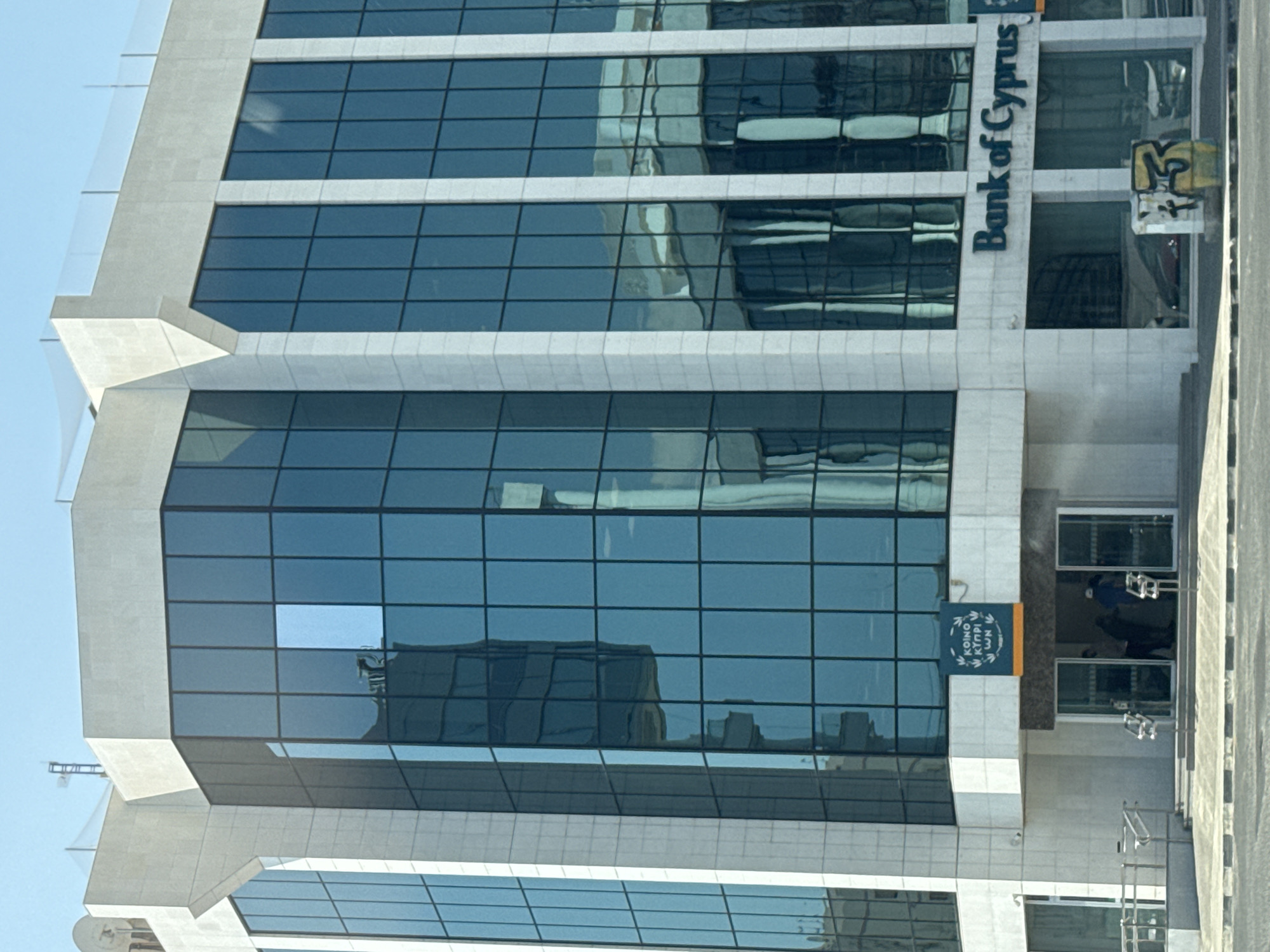 The image shows the Bank of Cyprus building located in Limassol, Cyprus. The building is a modern, multi-story structure with a facade of light-colored stone and dark blue tinted glass windows. The windows reflect the surrounding environment, showing distorted images of other buildings and the sky. There is a sign with the Bank of Cyprus logo and name displayed on the front of the building, to the right of the main entrance. The main entrance is recessed and features glass doors. Two individuals are visible entering or exiting the building. In front of the building, there is a sidewalk, a street, and a small concrete structure with graffiti on it. The overall scene depicts a typical urban commercial environment on a clear day.