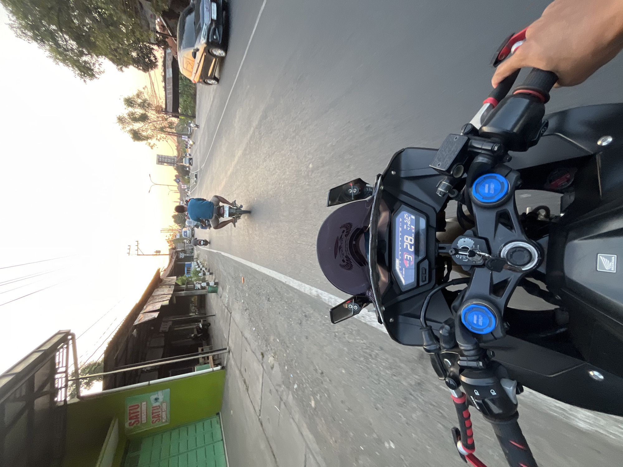 The image is a first-person perspective of someone riding a black motorcycle through a city street in Sengonbugel, Indonesia. The motorcycle's dashboard displays "3:28". On the left side of the street, there are green buildings with signs that read "SATU" and "SATU". Further down the street, several other motorcycles and a black car are visible. The sky is bright with a hint of sunset. The road is paved and appears to be a typical urban street.