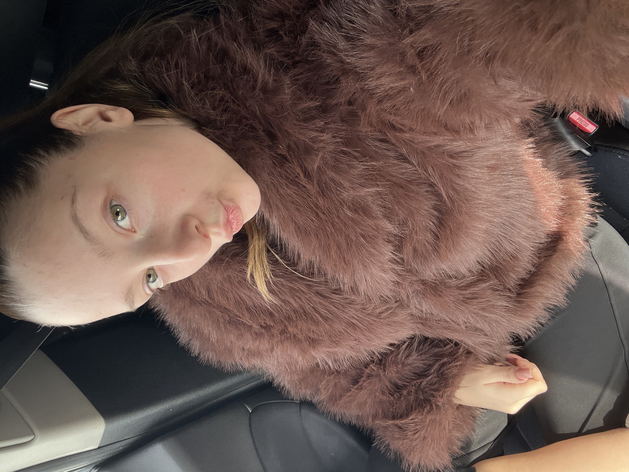 A young woman is angled at 90 degrees, appearing to be in a car, judging by the seat belt buckle, car seat and dashboard trim visible. She is wearing a textured, brown fluffy garment, possibly a jacket or wrap. Her skin tone is fair, and she has light brown hair. The setting does not immediately provide clues to suggest the location of Varna, Bulgaria.