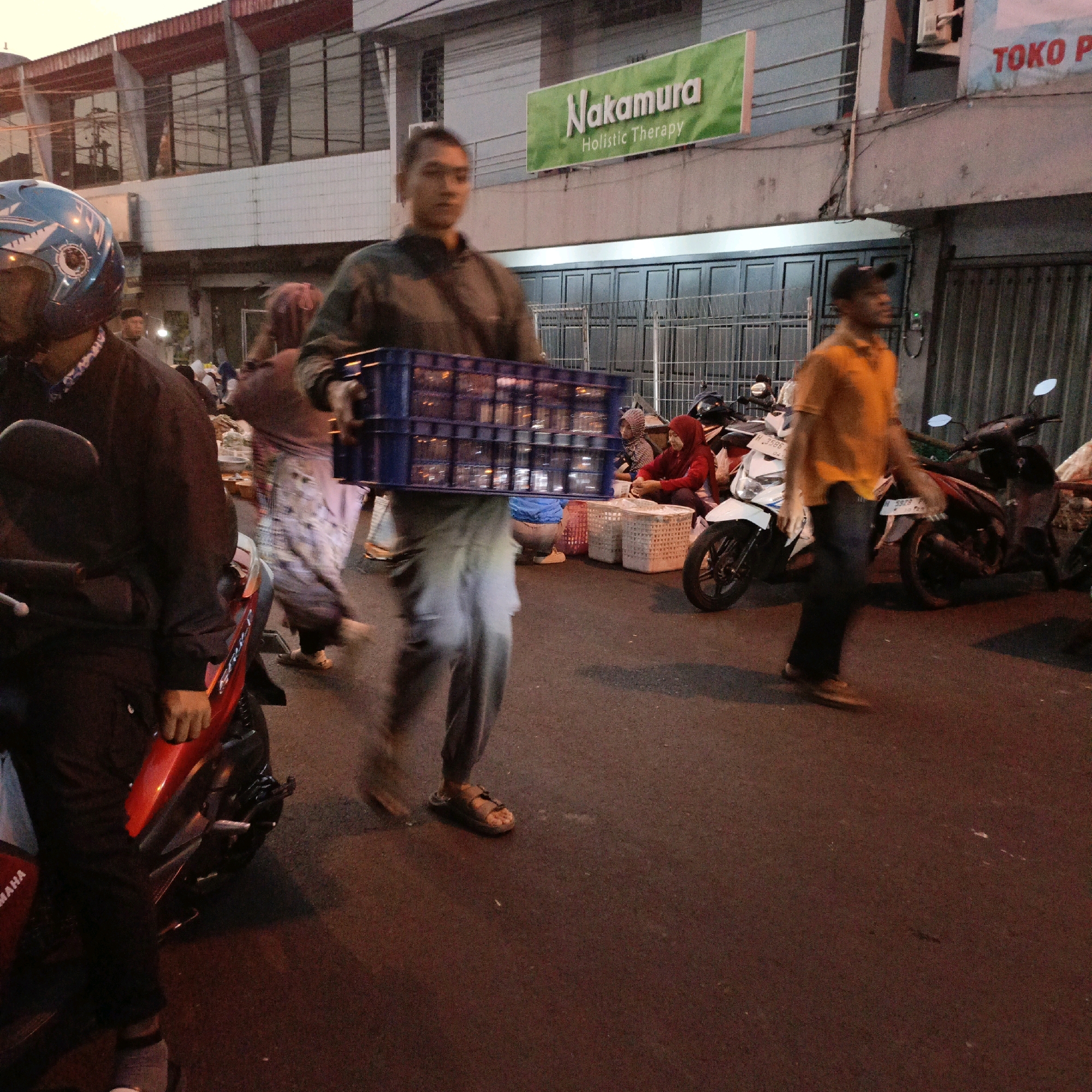 In Salatiga, Indonesia, a street scene unfolds as several individuals are captured in motion. In the foreground, a person wearing a blue helmet with a shark design on a motorcycle is partially visible. To the left, a man in a green shirt and grey pants carries a blue plastic crate filled with clear containers, walking on a paved road. Behind him, another person, possibly a woman in a headscarf, is seen walking away. In the background, a sign for "Nakamura Holistic Therapy" hangs above a building with large windows. Further to the right, a man in an orange shirt is walking past parked motorcycles. The environment suggests a market or bustling street with several scooters and some stalls visible, illuminated by the warm, low light of dusk.